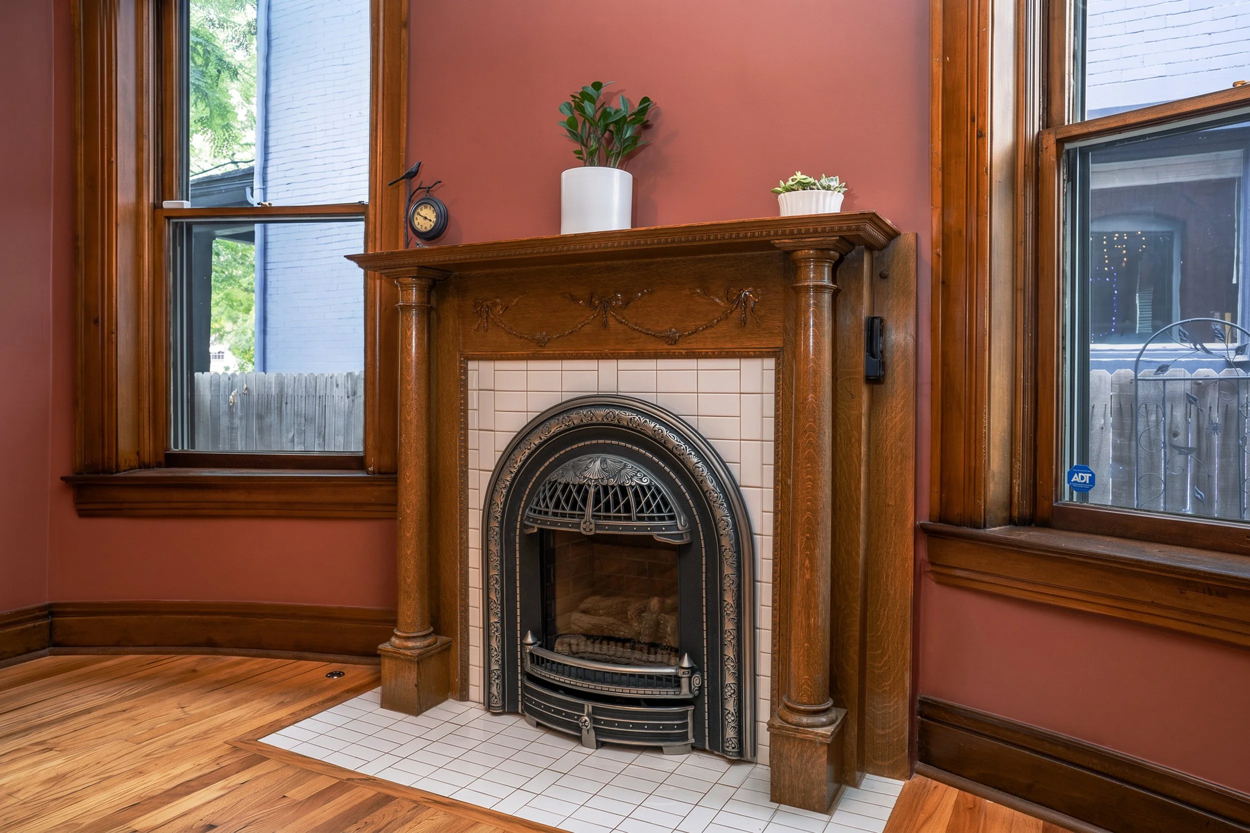 A fireplace with an ornate black and gold metal surround, white tile, and a wooden mantel with decorative carvings. There are two windows on either side of the fireplace, framed with wood, showing trees and a fence outside. The walls are painted a reddish color.