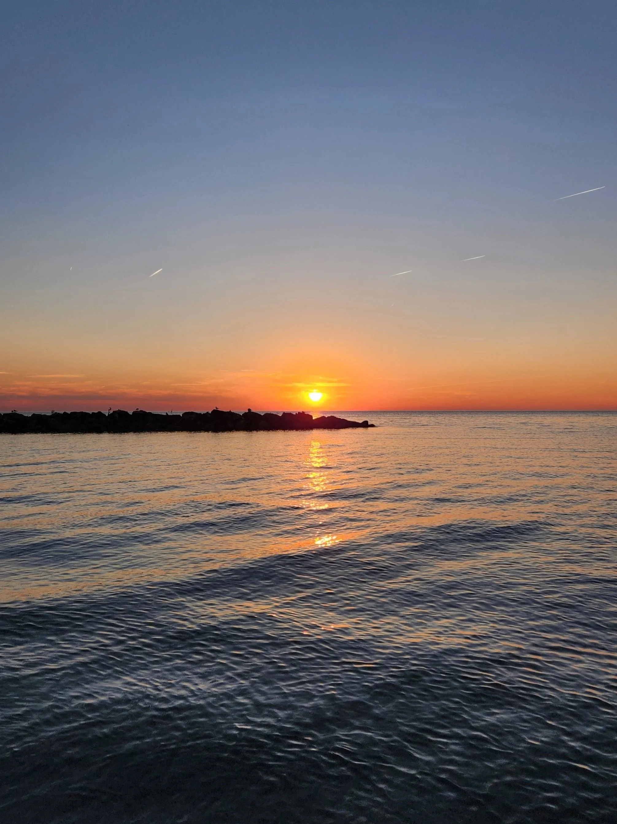A sunset over a body of water with a rocky shoreline silhouette in the foreground and a clear sky with a few visible contrails.