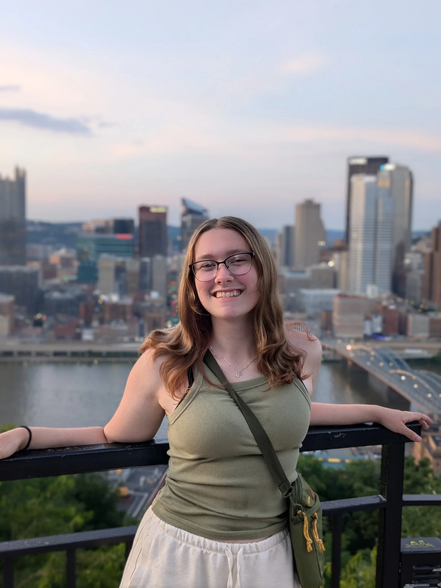 A smiling young woman with glasses, wearing a green tank top and white pants, standing on a balcony overlooking a city skyline in the evening.