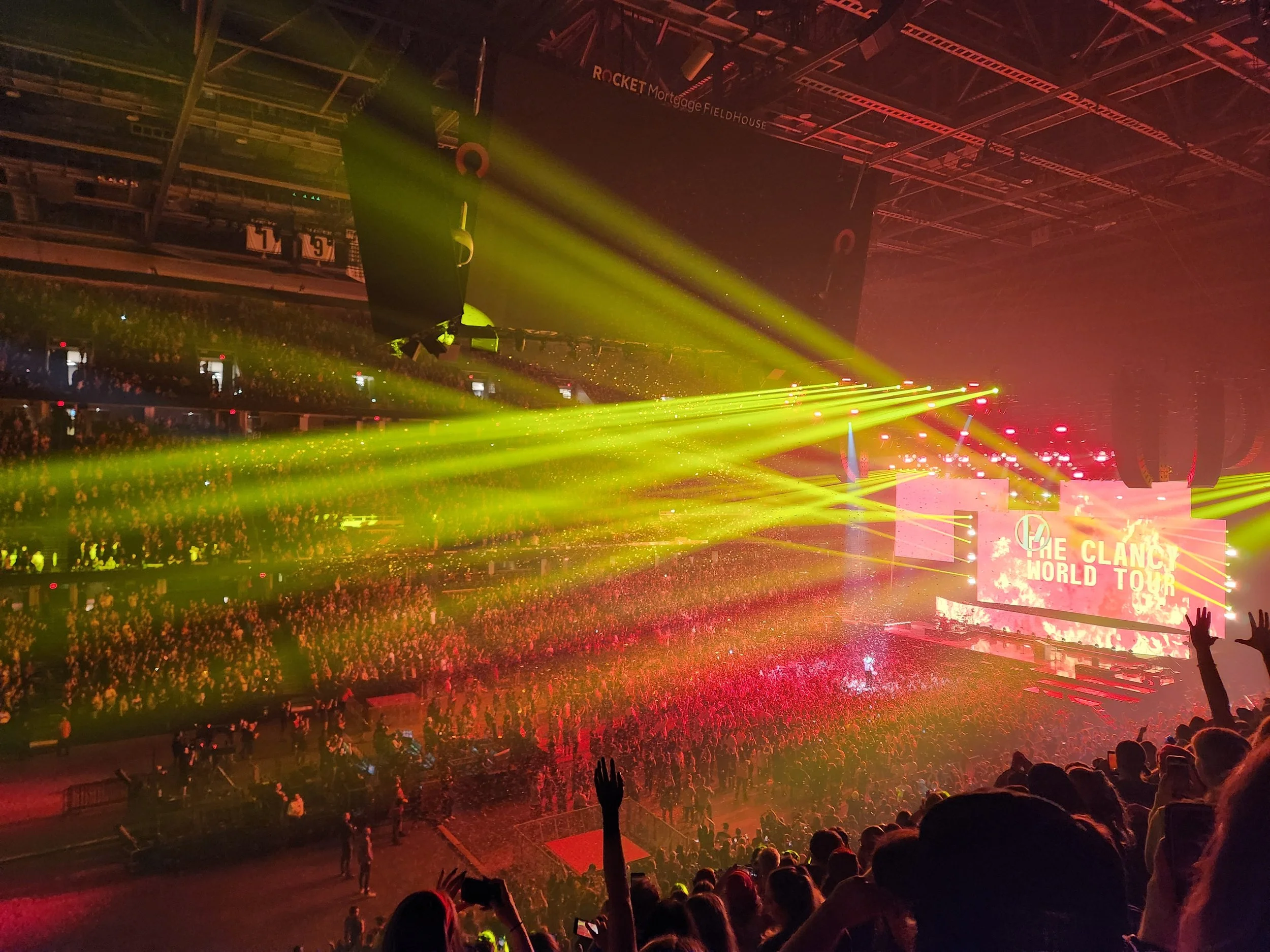 An indoor concert scene with yellow stage lights, a large crowd, and a screen displaying 'The Clancy World Tour.'