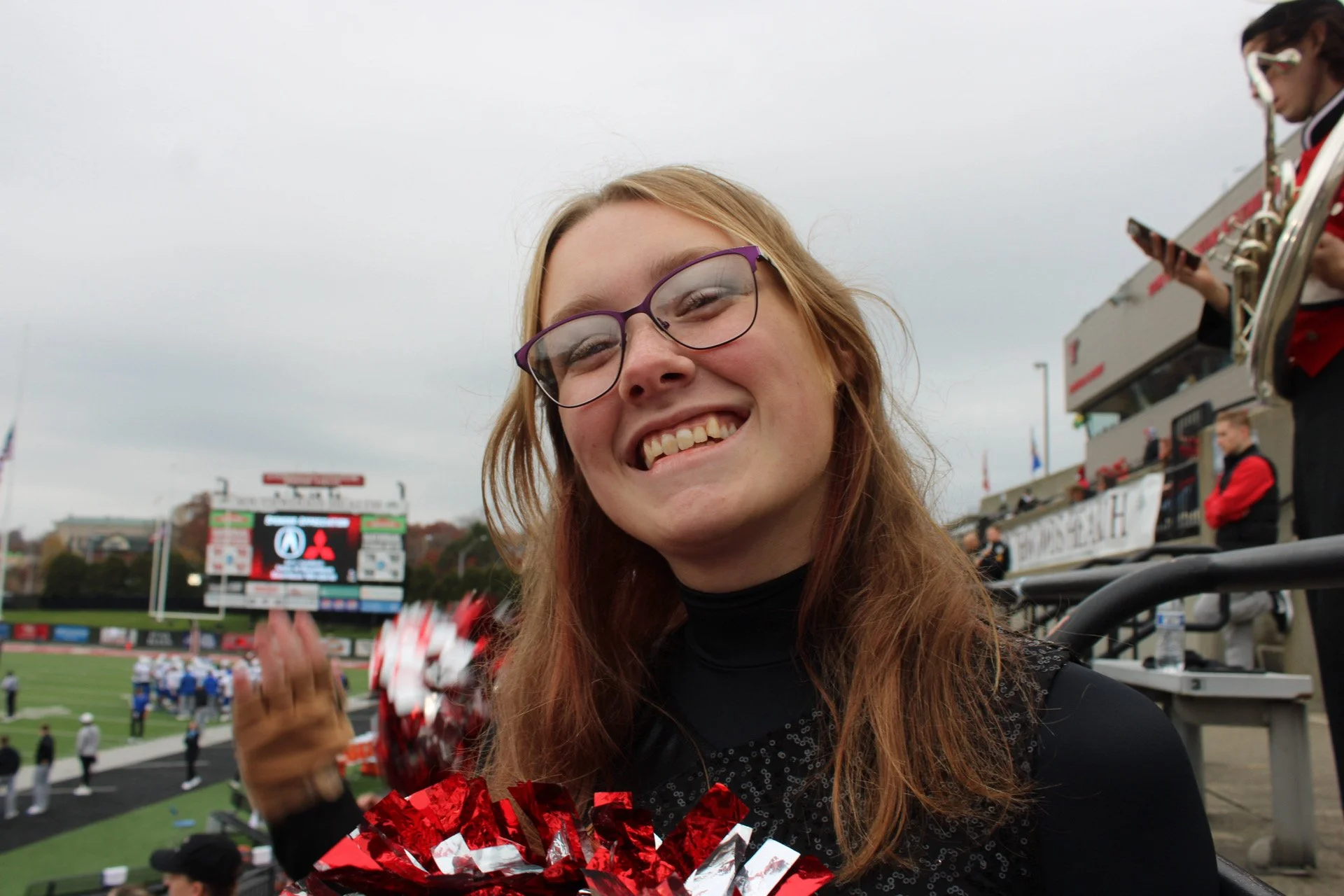 A young woman with long red hair and glasses smiling at a sports stadium, holding red and white pom-poms, with a scoreboard and other people in the background.