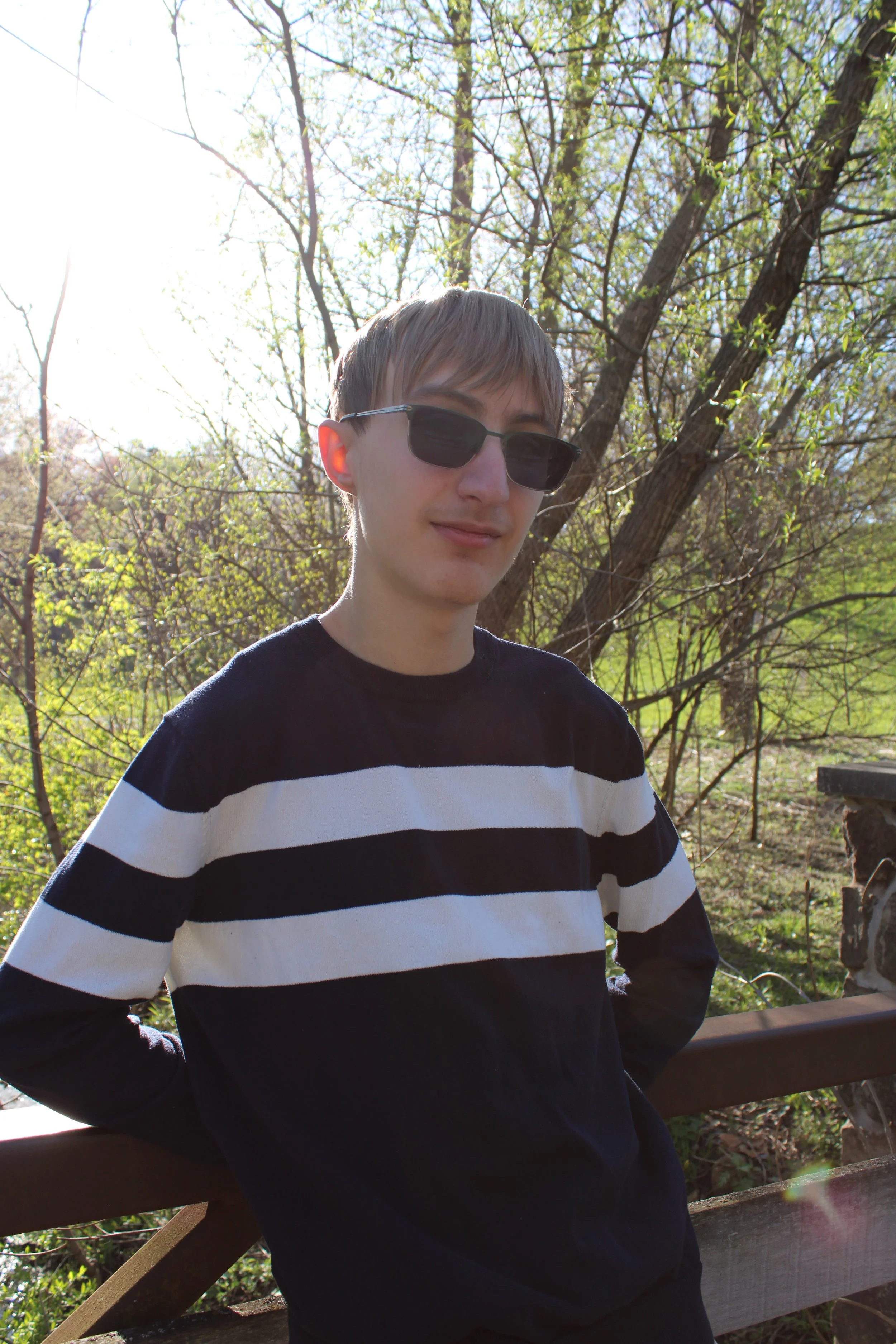 A young man wearing sunglasses and a black and white striped shirt, standing outdoors in front of trees and sunlight, with a wooden railing in front.