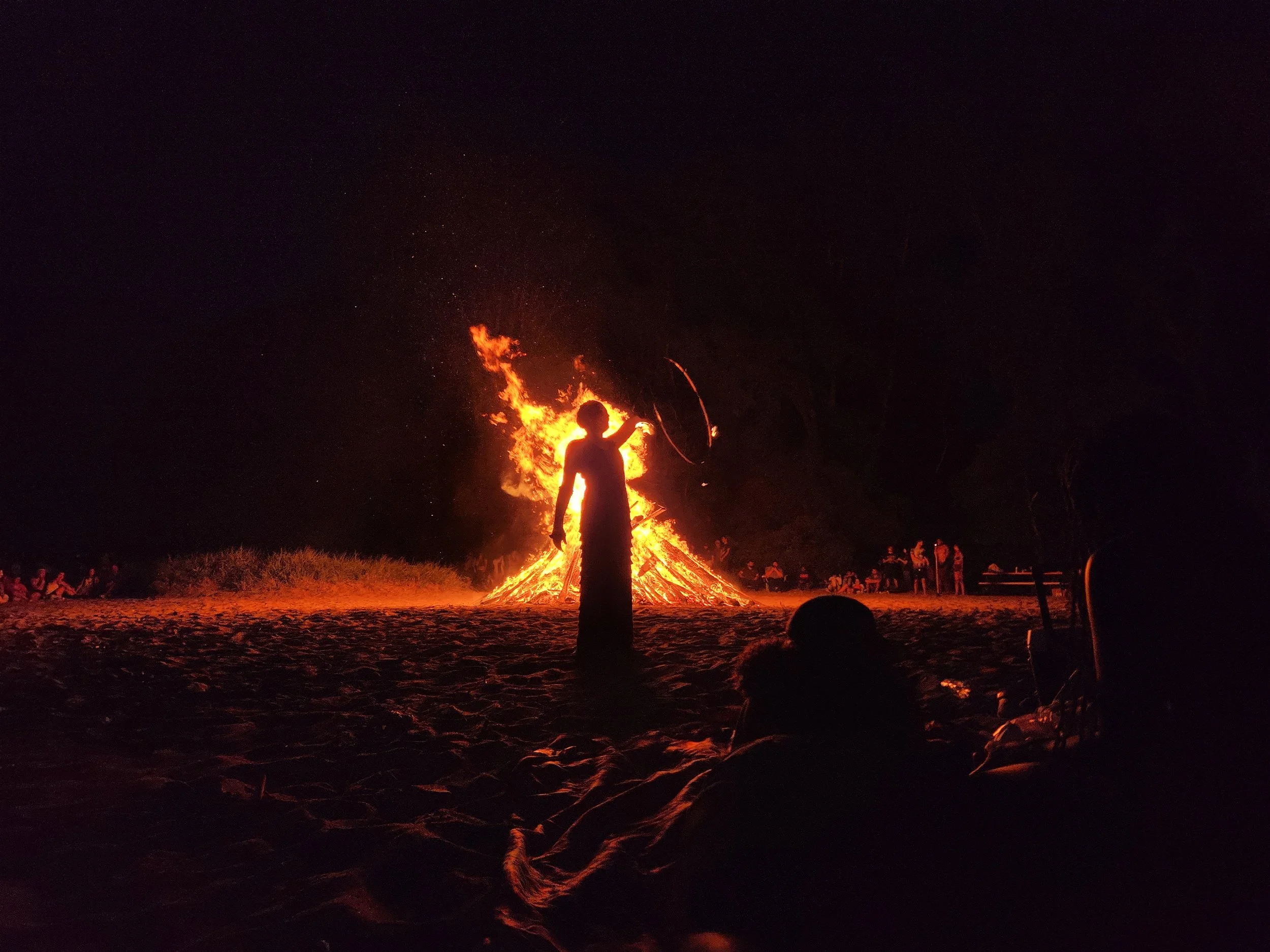 Person standing near a large bonfire at night, with people in the background.