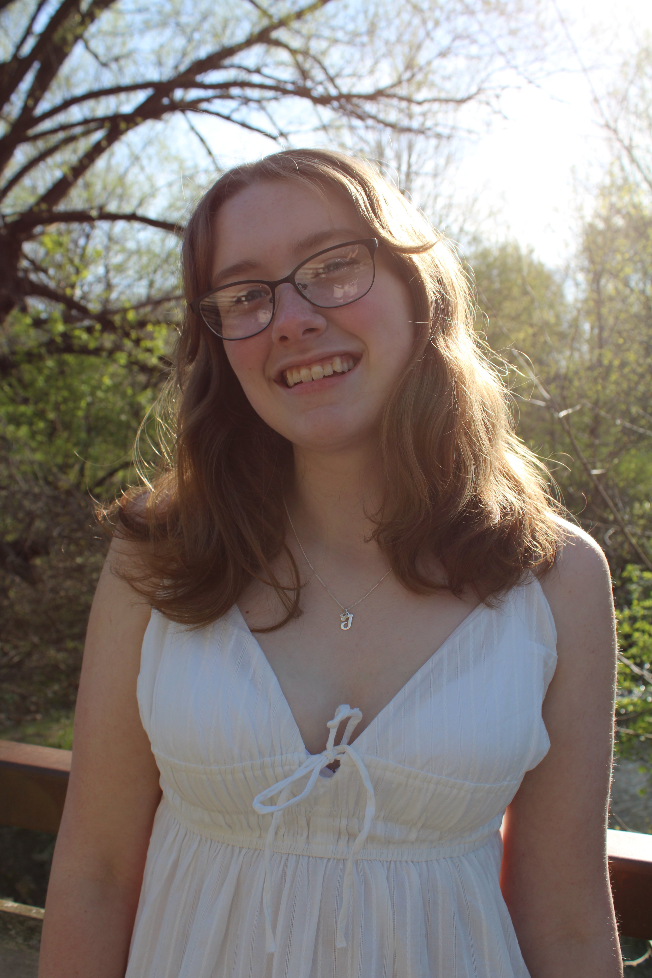 A young woman with wavy brown hair and glasses smiling outdoors on a sunny day, standing in a garden area with trees and foliage in the background.