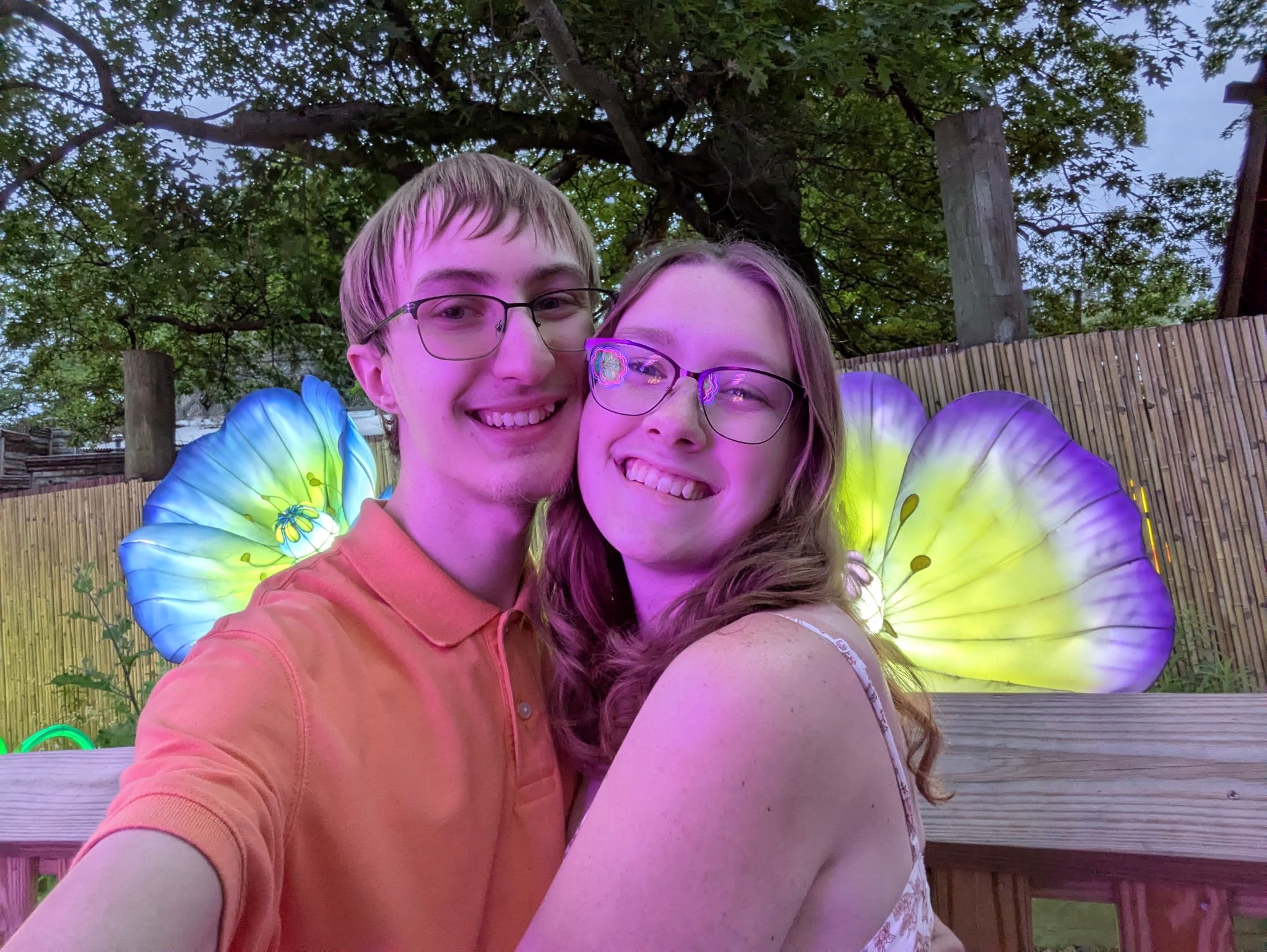 Two young people smiling and taking a selfie outdoors during twilight, with a wooden fence and trees in the background, and colorful glowing decorations around them.