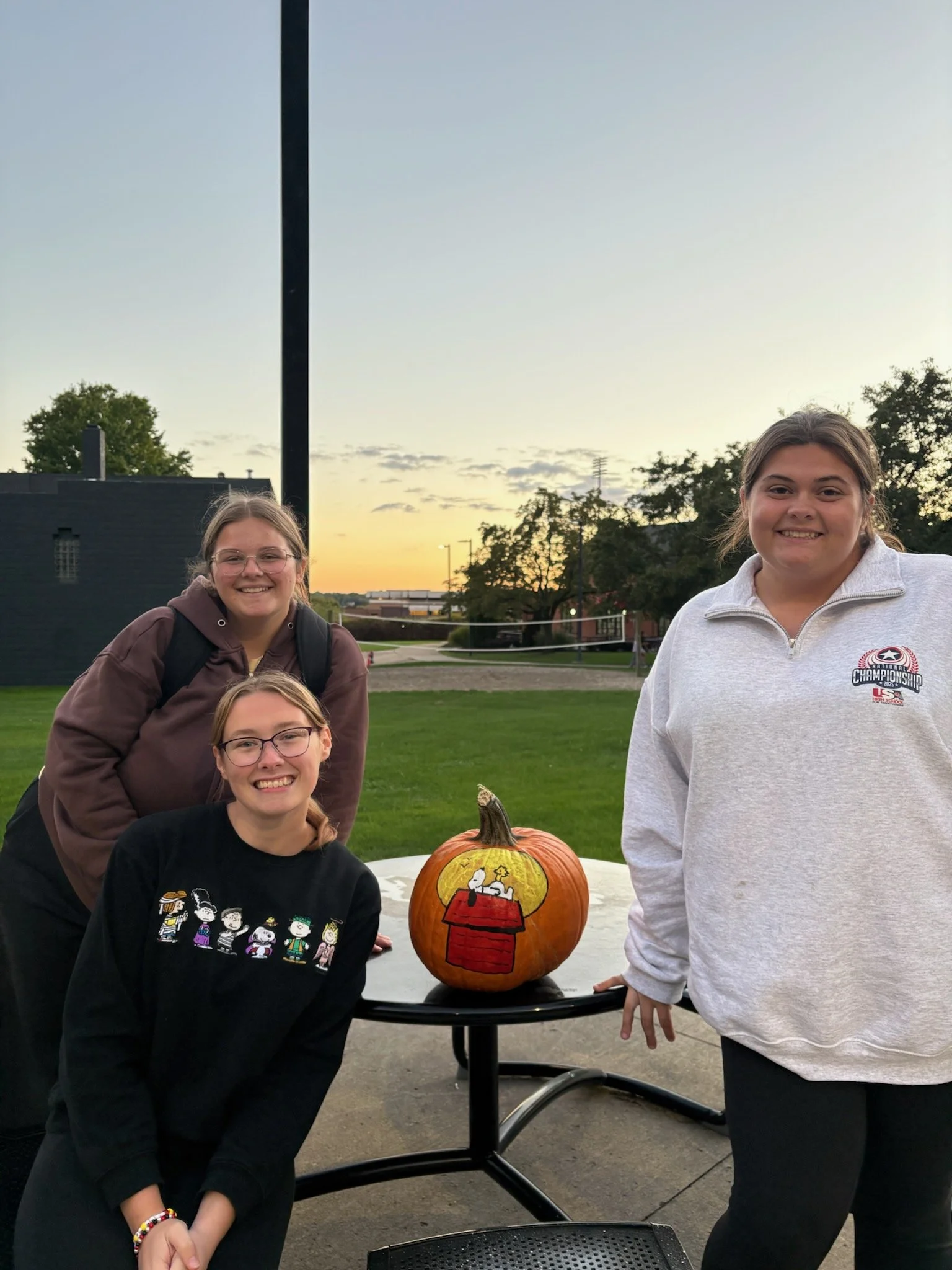 Four girls posing outdoors near a decorated pumpkin on a small table during sunset. Two girls are standing behind, one kneeling, and one sitting in front, all smiling.