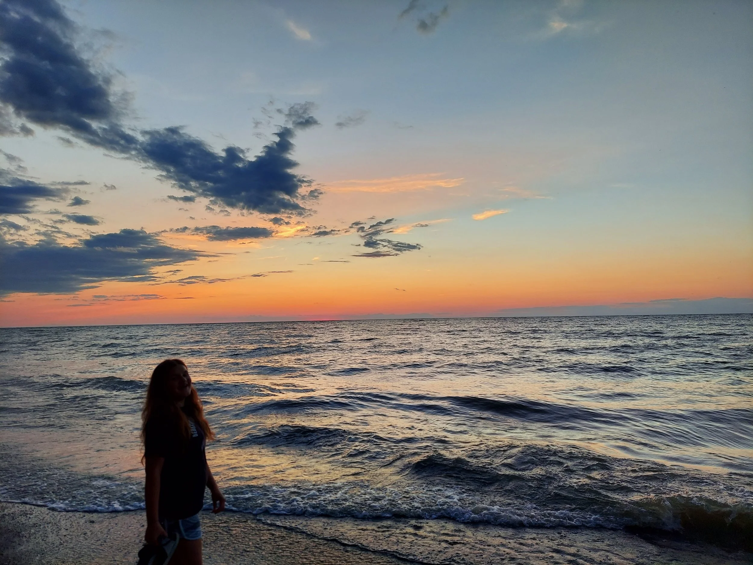 Person standing on a beach at sunset with colorful sky and waves.