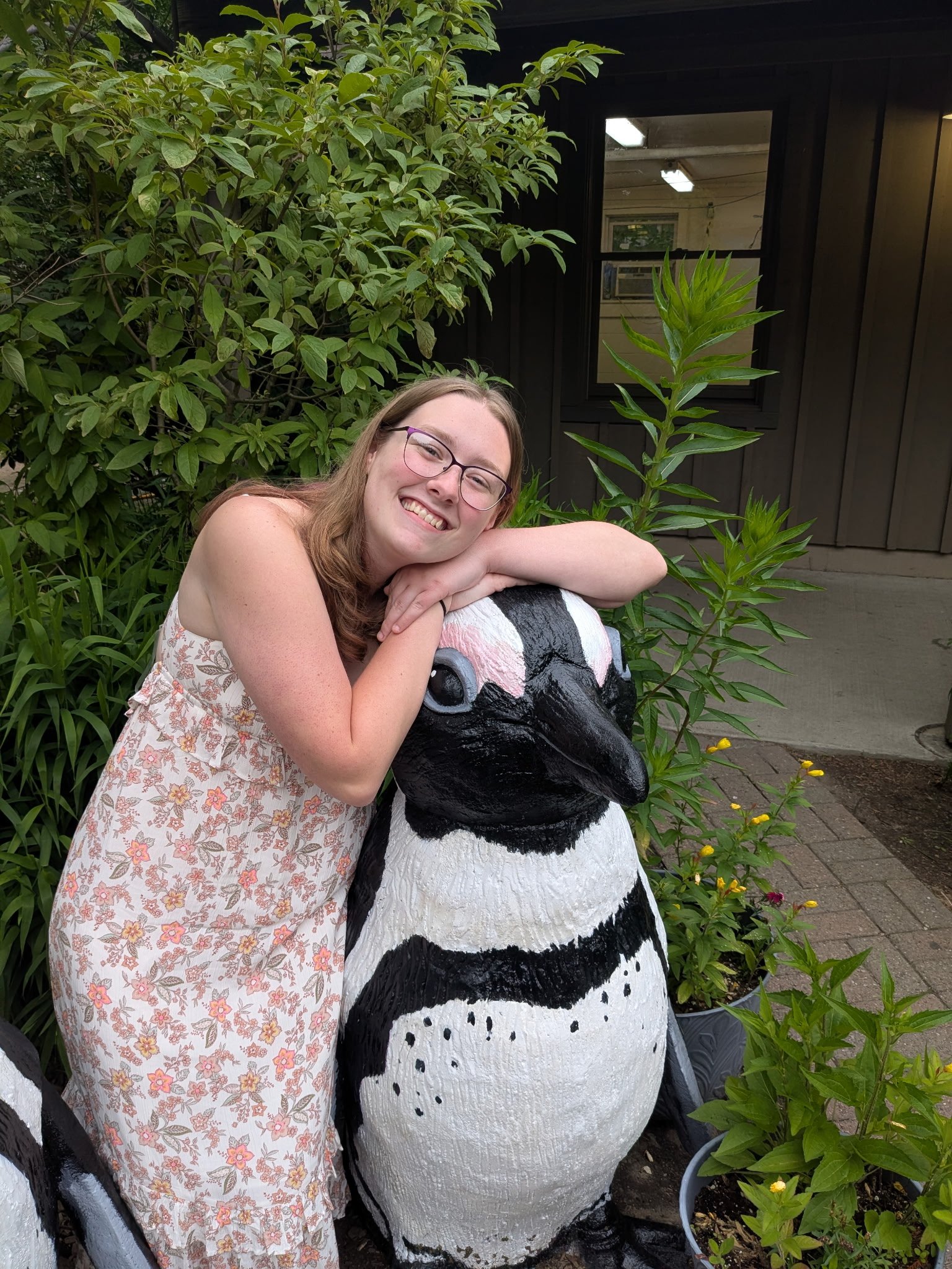 A smiling young woman with glasses, wearing a floral dress, leaning on a large penguin sculpture outdoors surrounded by greenery.