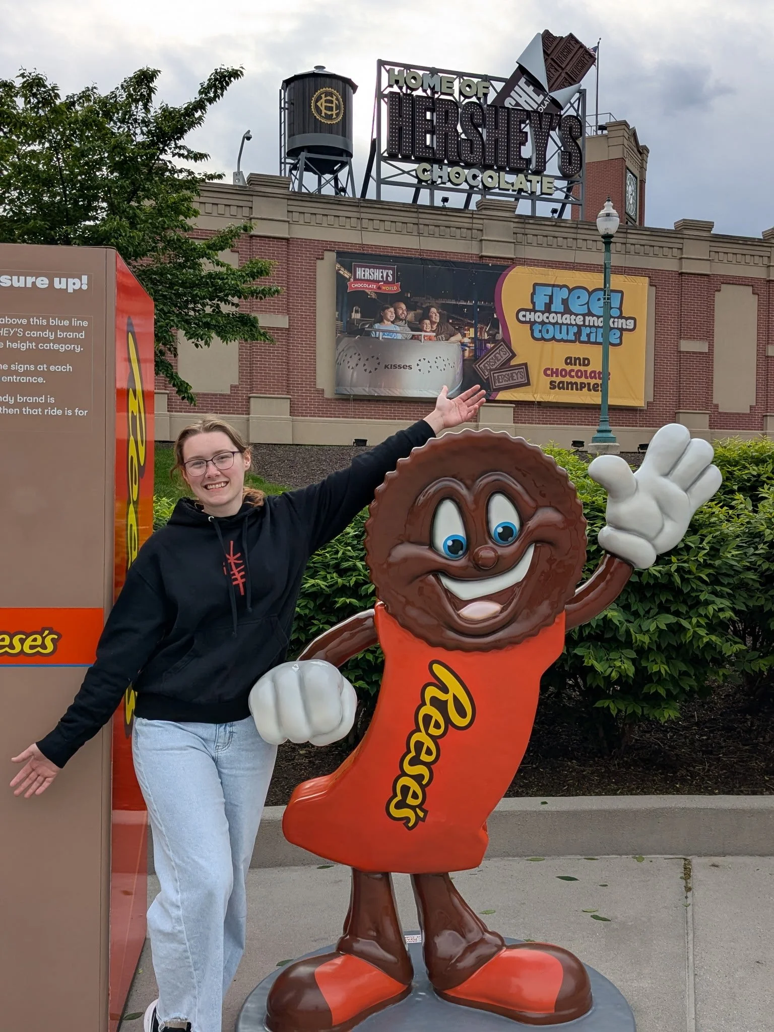 A young woman wearing glasses and a black hoodie, smiling and gesturing with her arms, standing next to a large cartoonish chocolate character mascot with a smiling face, big eyes, and white gloves, at a theme park