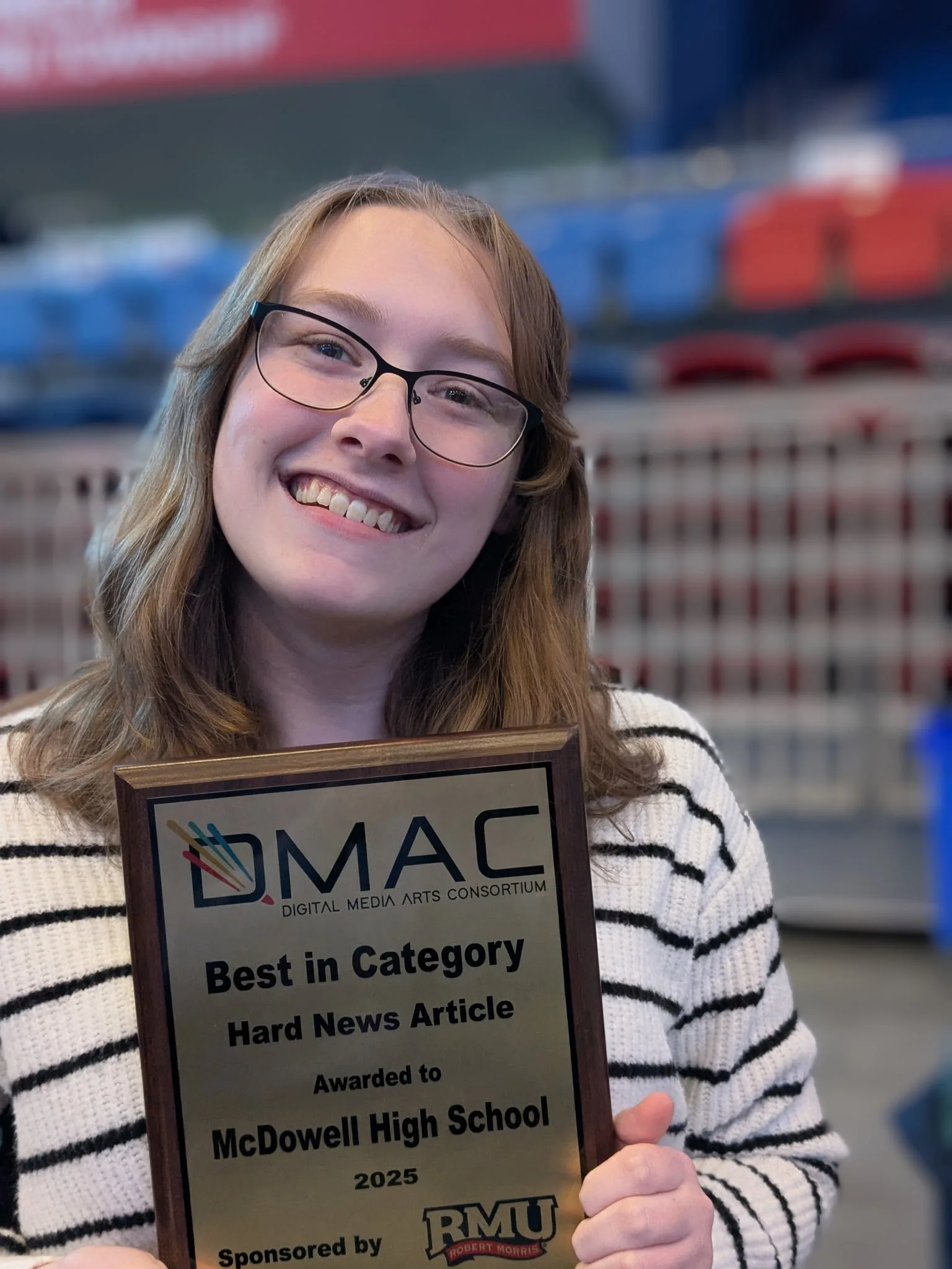 A young woman with glasses and long hair smiling, holding an award plaque for best in category in news writing.