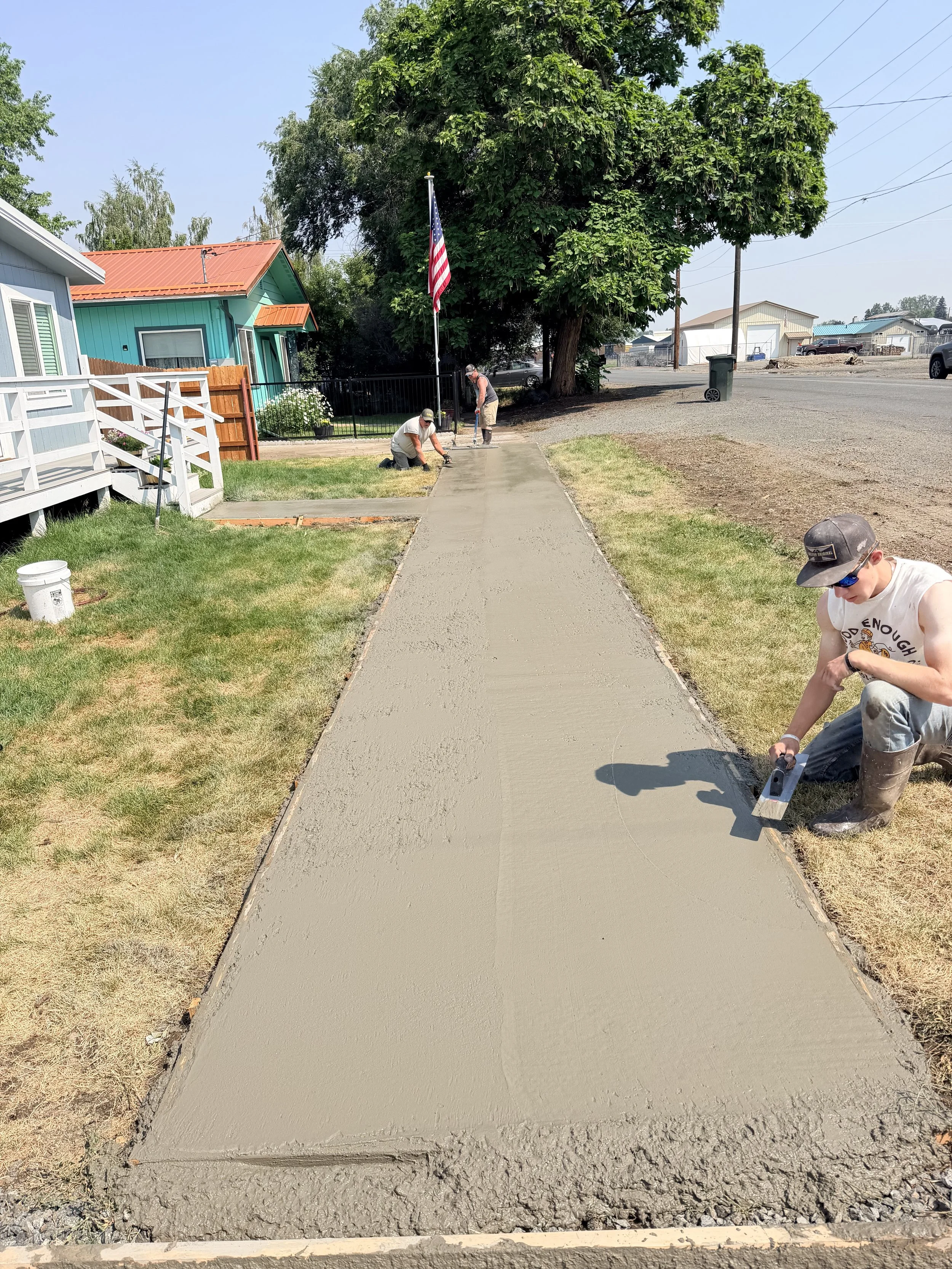 People are pouring and smoothing fresh concrete on a sidewalk in a residential neighborhood during daytime.