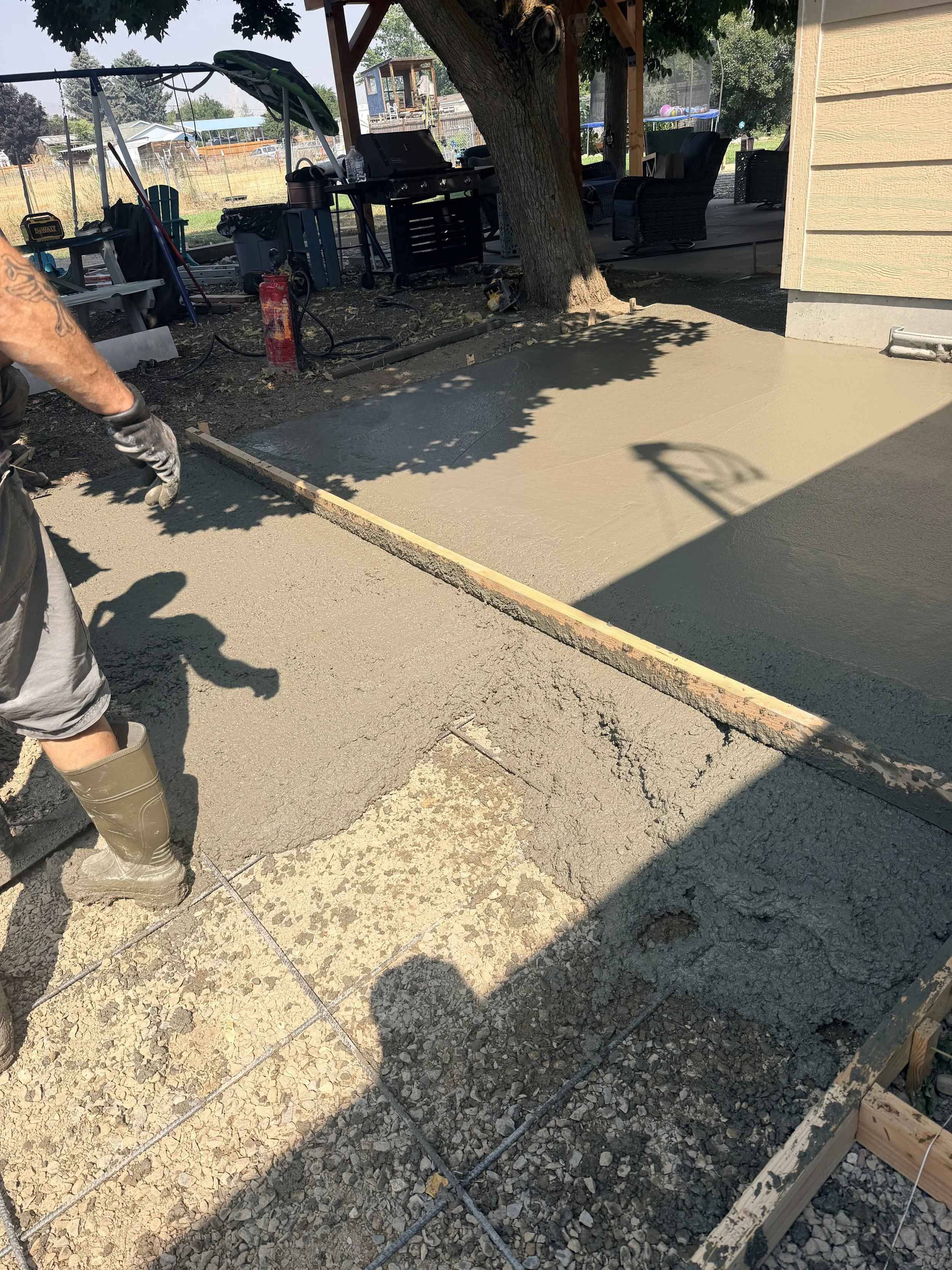 A person wearing rubber boots is pouring concrete for a patio or walkway, with some wooden forms and tools visible. A tree and a covered outdoor seating area are also in the background.