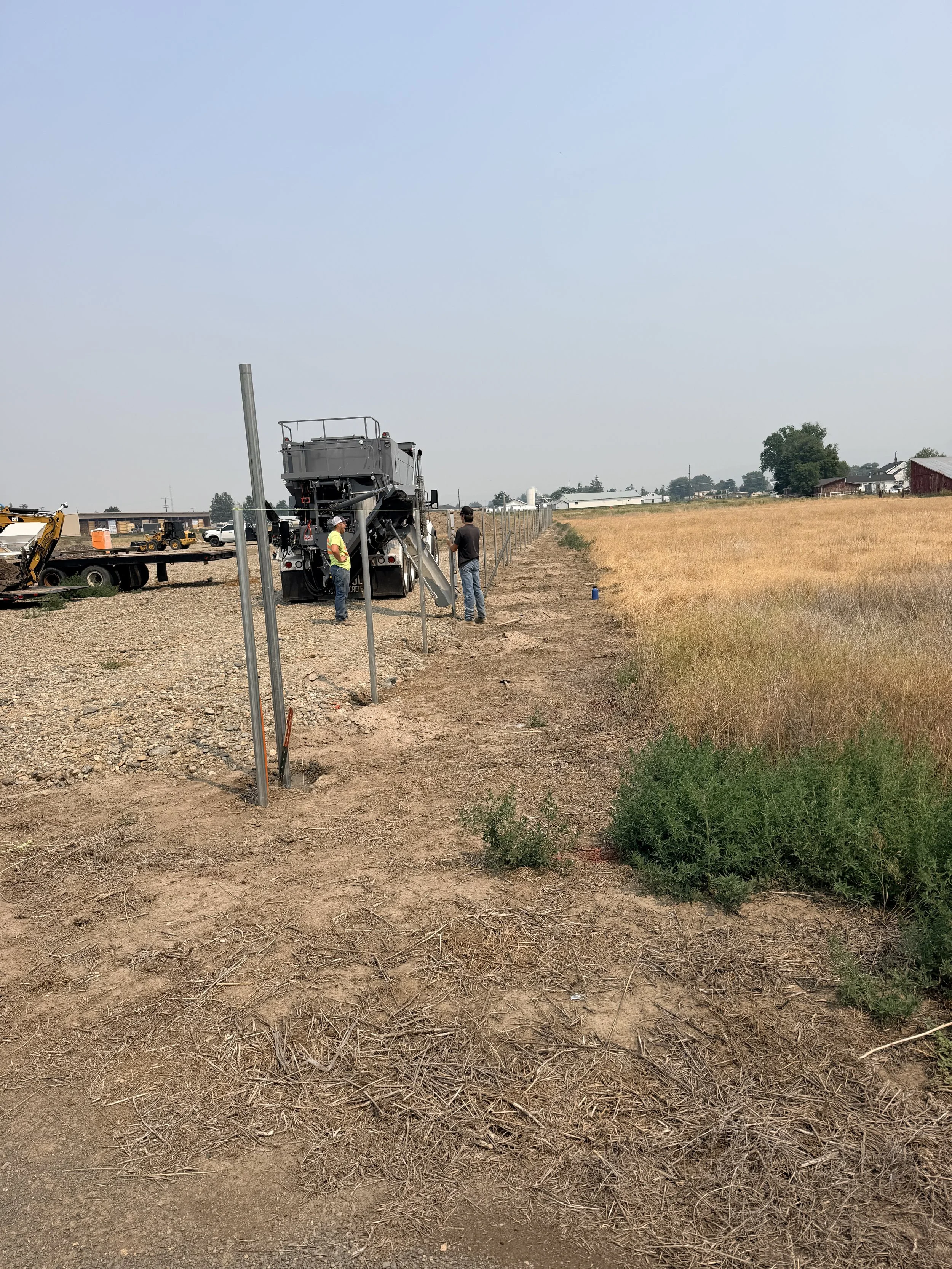 Construction workers and equipment set up fencing in a rural area with fields and small buildings in the background.