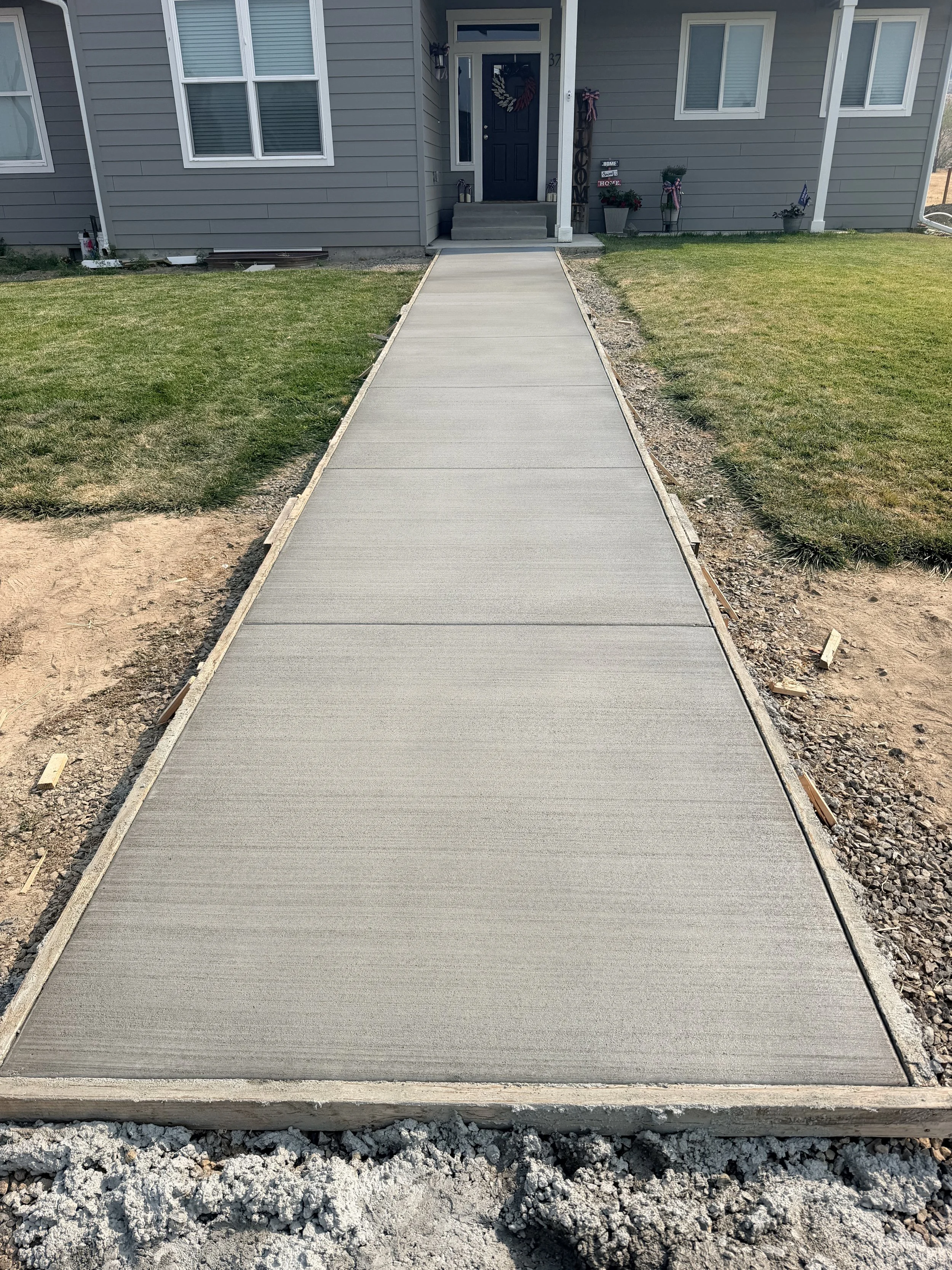 Freshly poured concrete sidewalk leading to the front porch of a house with gray siding and black door, surrounded by green lawn and landscaping.