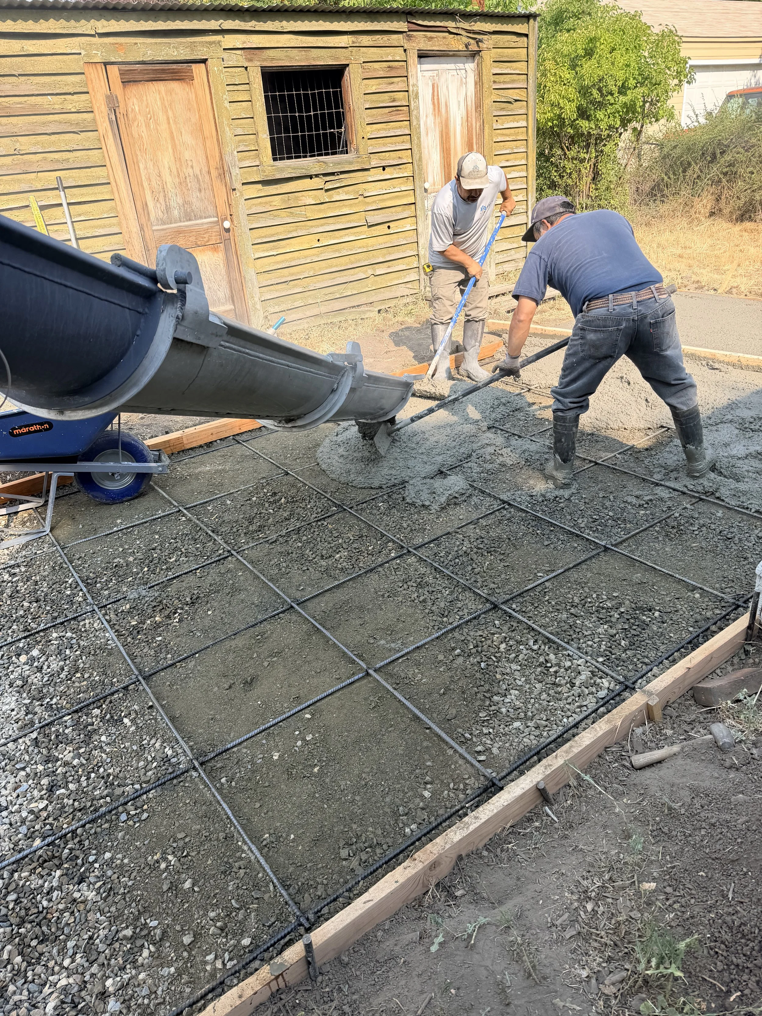 Two workers are pouring wet concrete into a rebar grid for a small concrete slab in a backyard, with a wooden shed in the background.