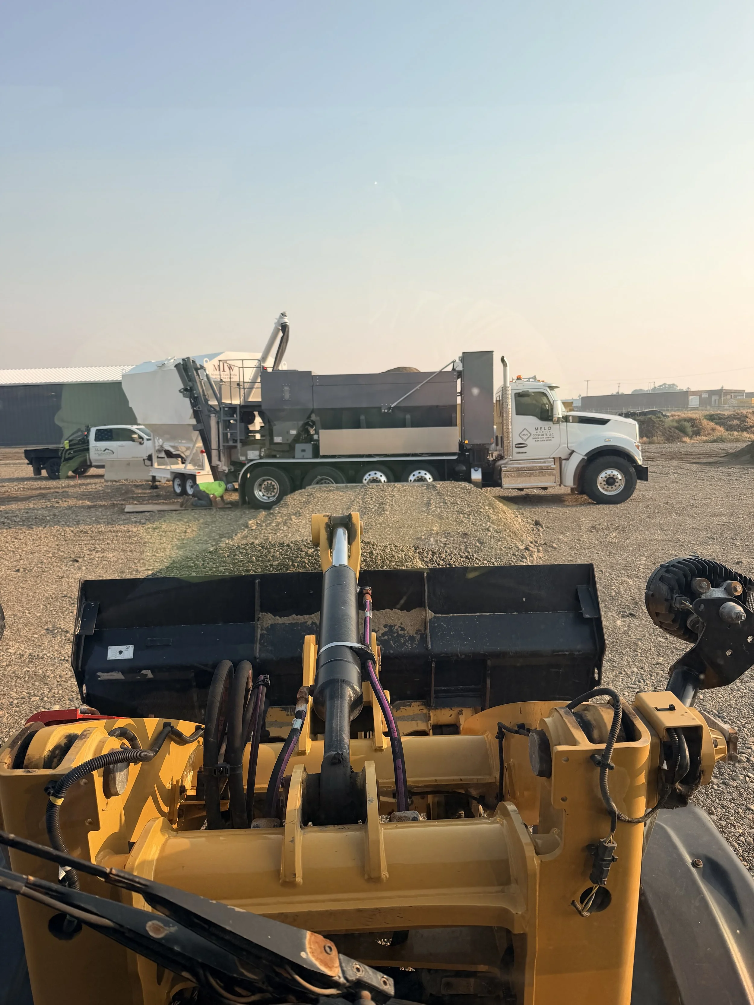 View from the operator's seat of a construction vehicle, showing a large, white and gray construction truck with equipment and a person working nearby, set in a gravel area with buildings in the background.