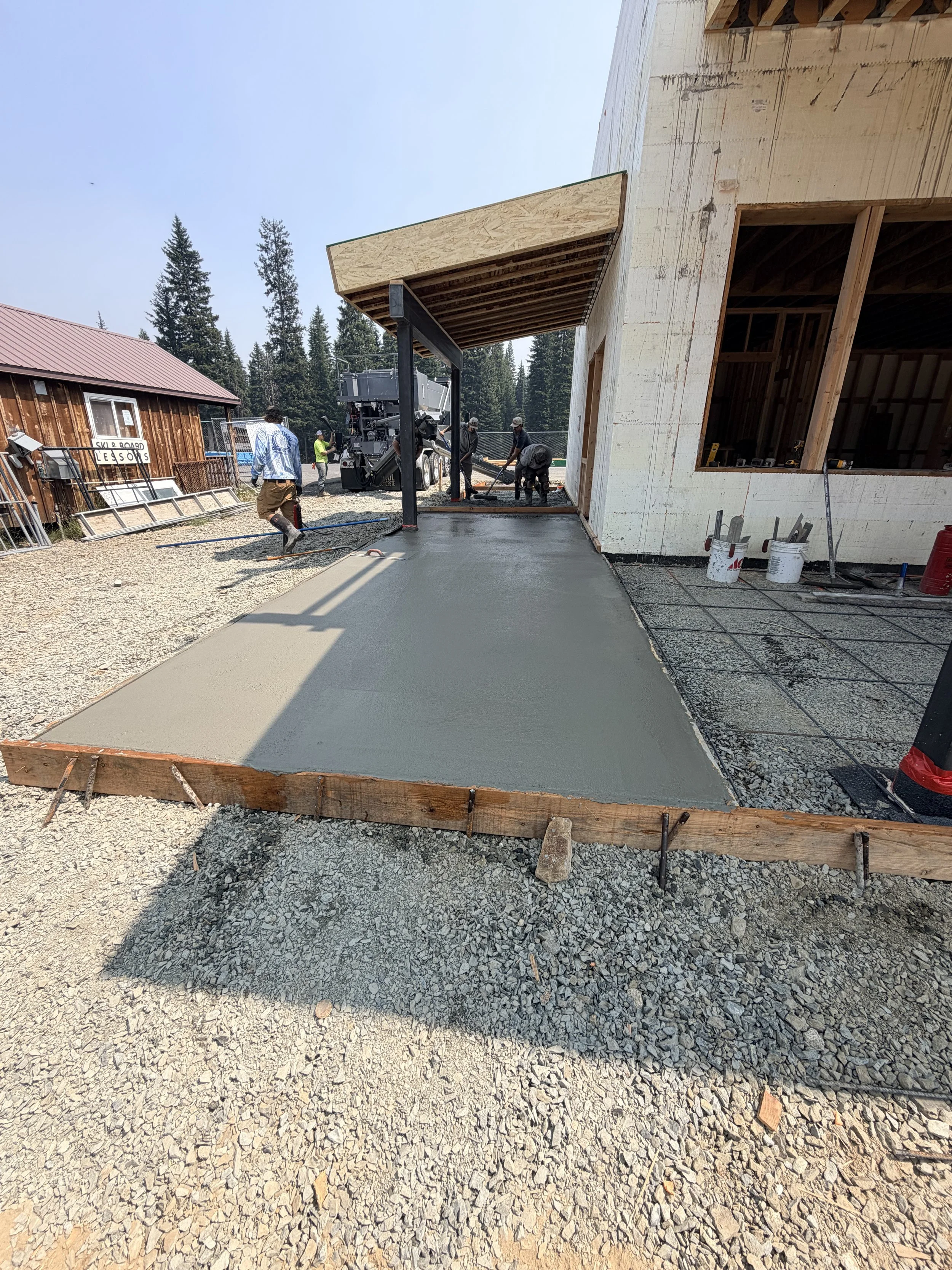 Construction workers pouring and leveling concrete for a slab in front of a partially built house, surrounded by gravel and construction tools.
