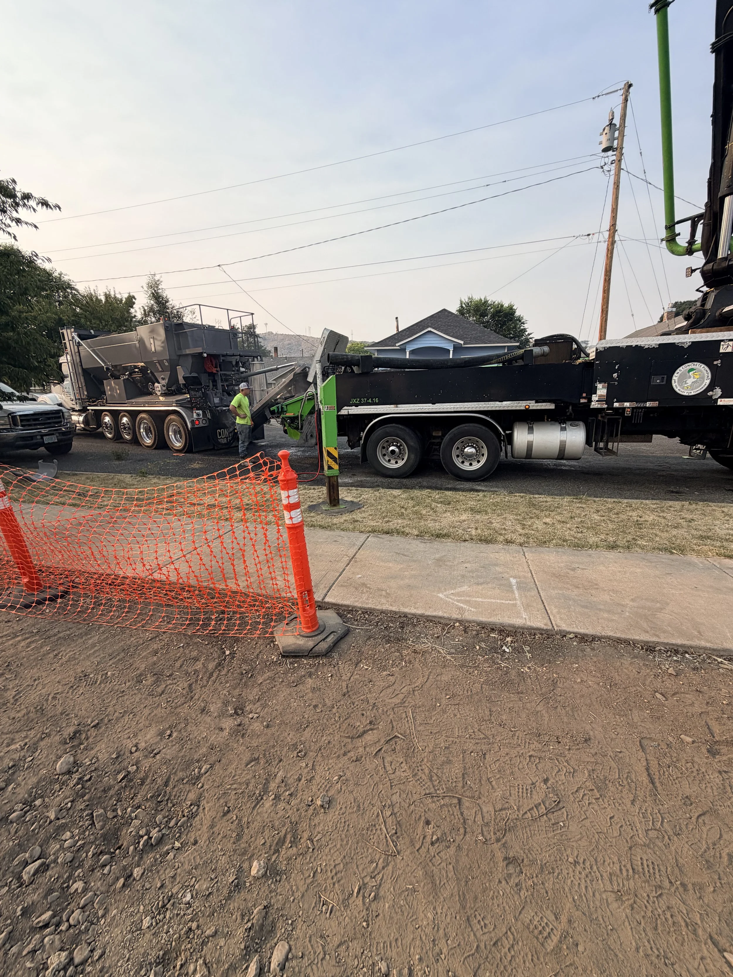 Construction site with heavy machinery, orange safety fence, and workers, and a residential house in the background.