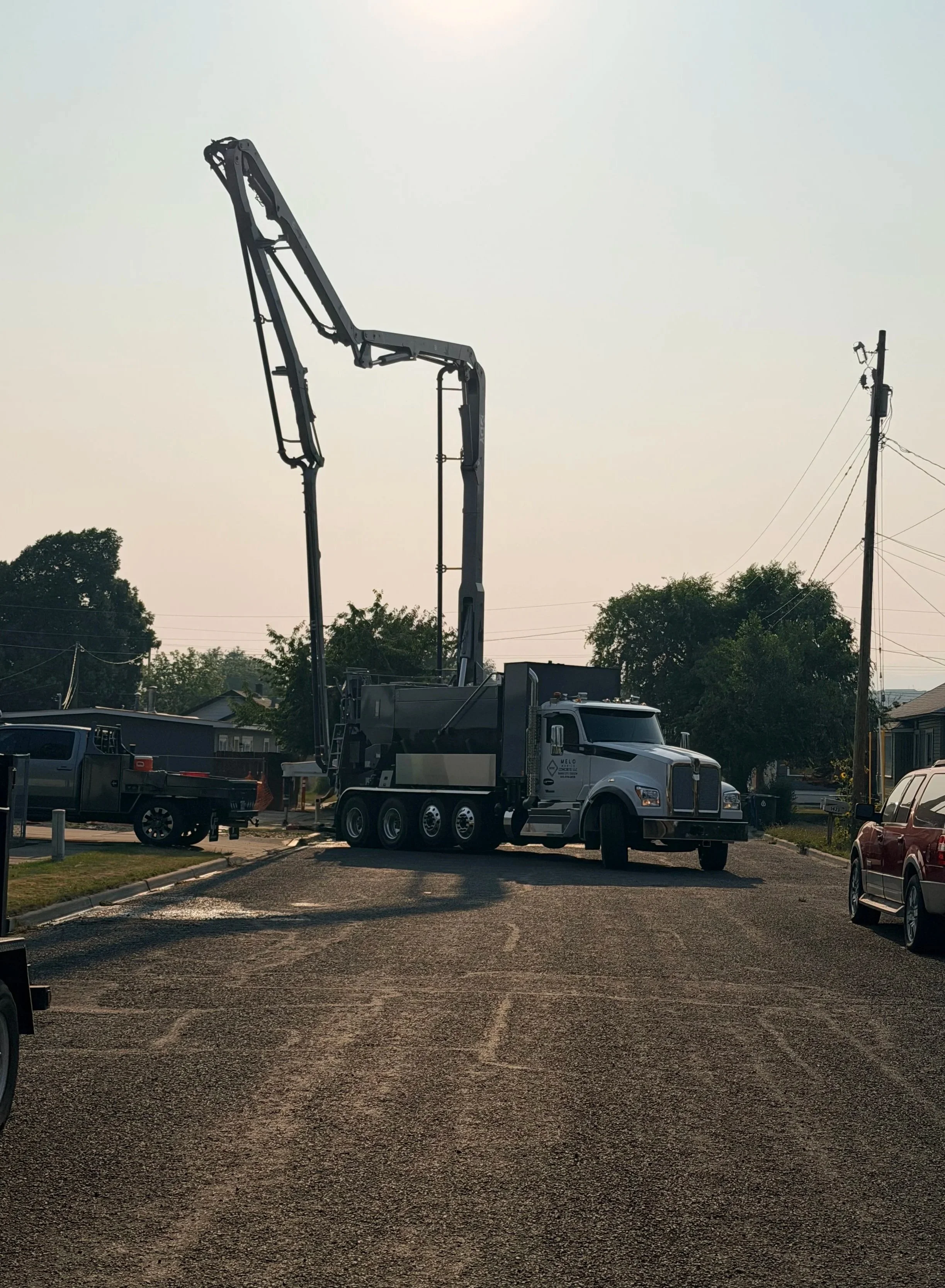 A large boom truck with a hydraulic arm is parked in a parking lot, with trees and utility poles in the background during daylight.