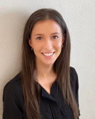A young woman with long brown hair, smiling, wearing a black top, standing against a beige wall.
