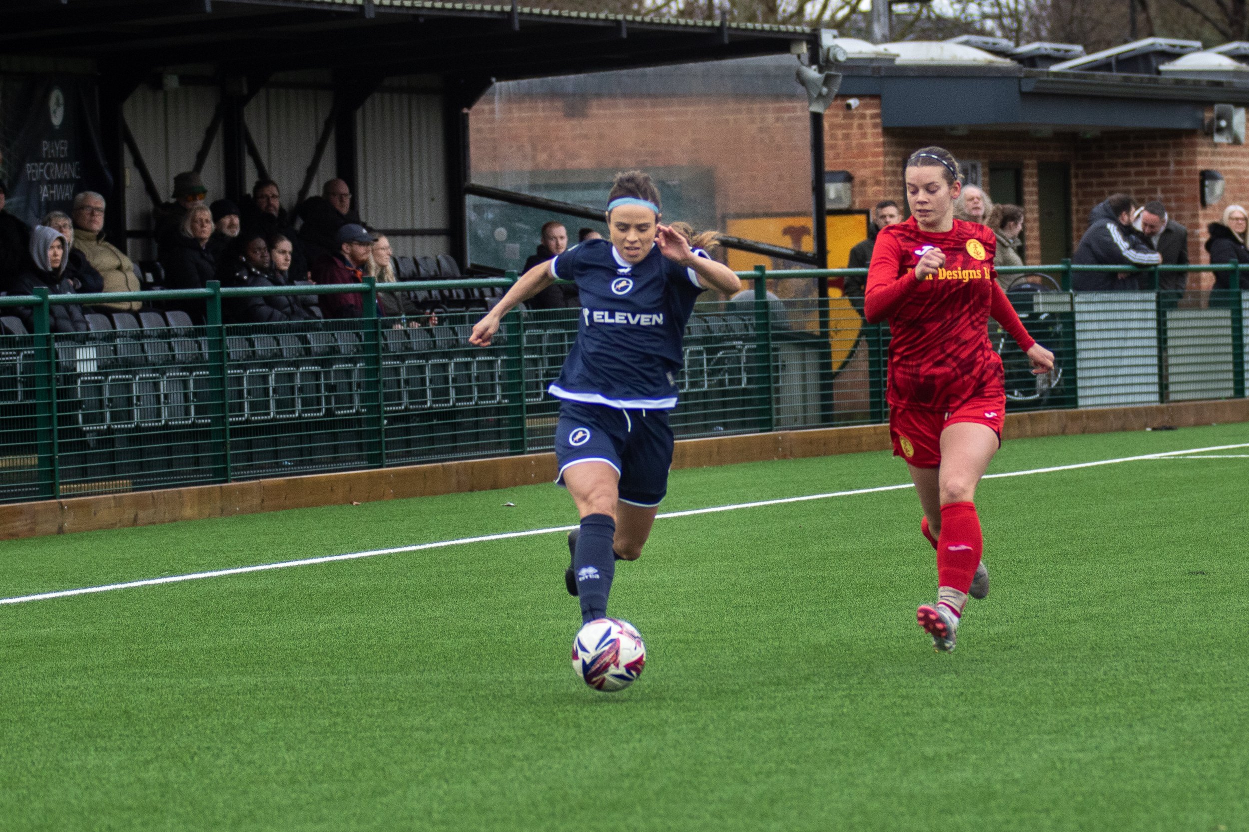Two female soccer players, one in a blue uniform and the other in a red uniform, running on a green field with a soccer ball at their feet during a match. Spectators are seated on benches and standing behind a green fence in the background.