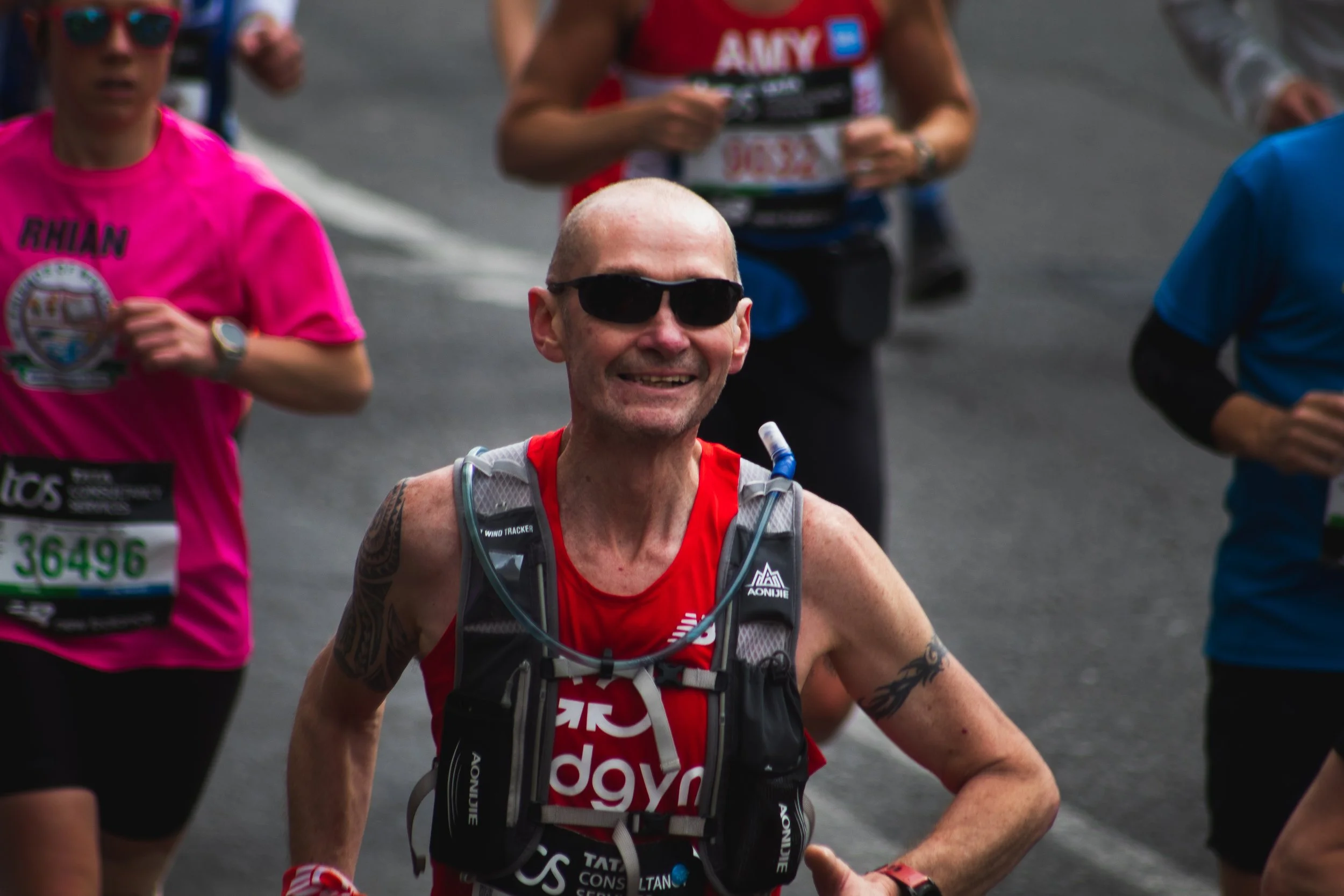 A man with a shaved head and sunglasses running in a race, wearing a red athletic shirt and a hydration vest. Several other runners are visible in the background, some wearing pink, blue, and red shirts.