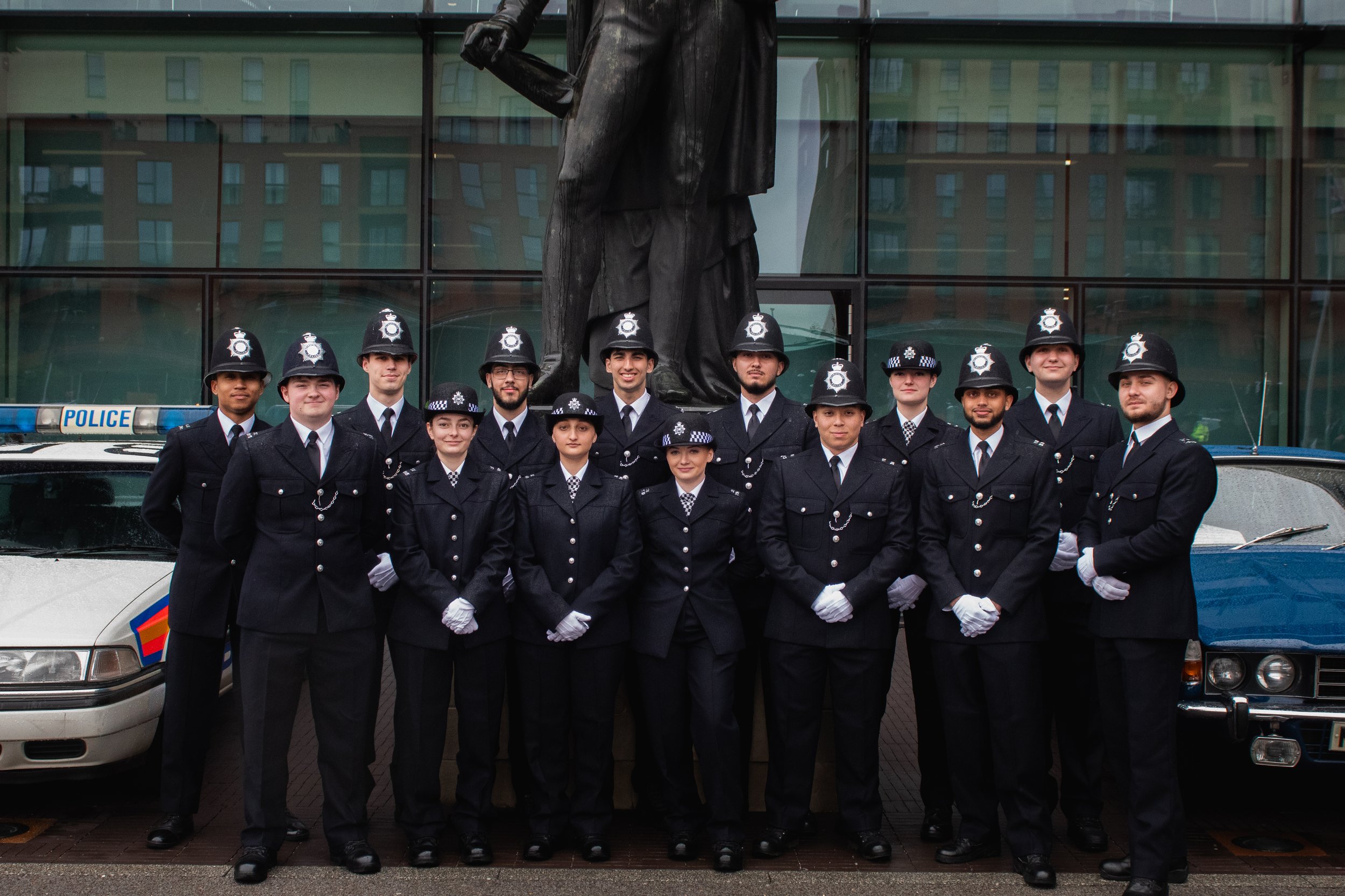 A group of young police officers in uniform standing in front of a police vehicle and a large building with reflections in the windows.