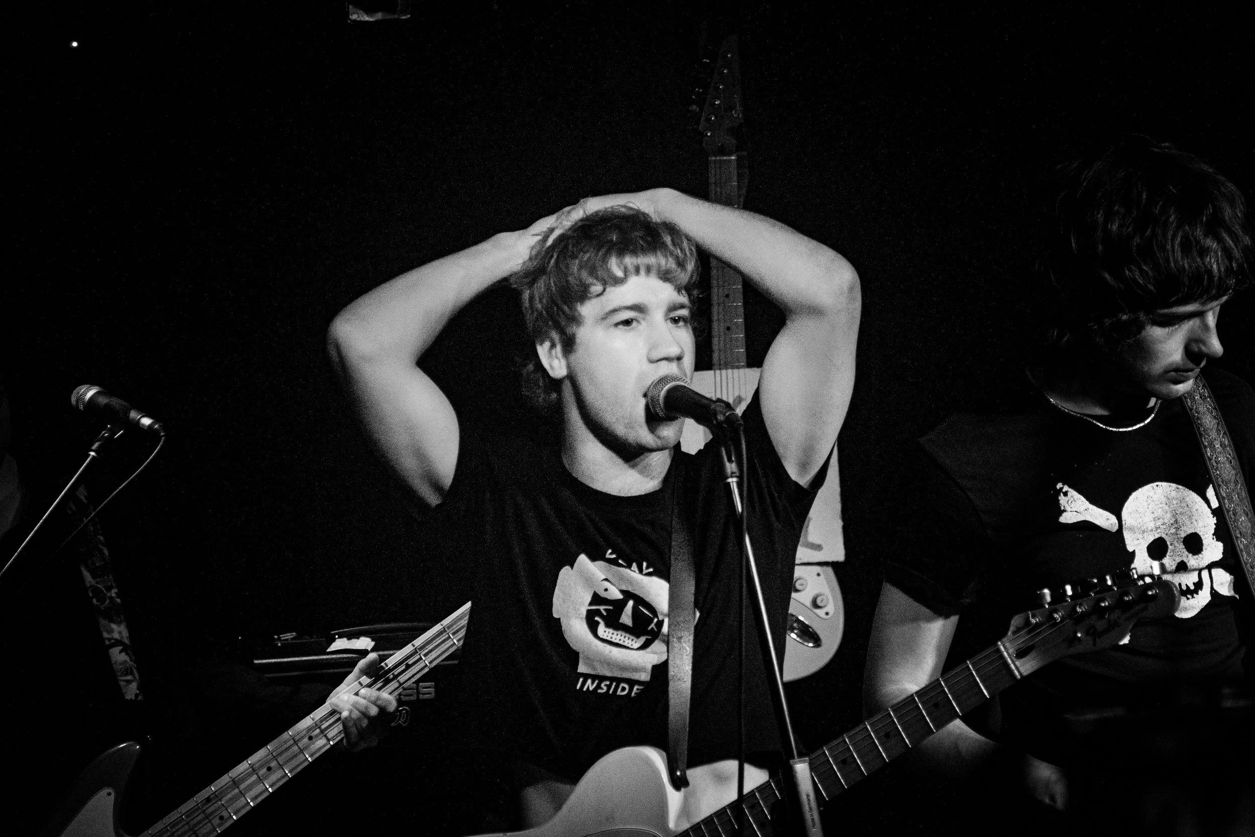 Black and white photo of two young men performing music. The man on the left is singing into a microphone, with his hands on his head, and holding an electric bass guitar. The man on the right is playing an acoustic guitar, wearing a shirt with a sku