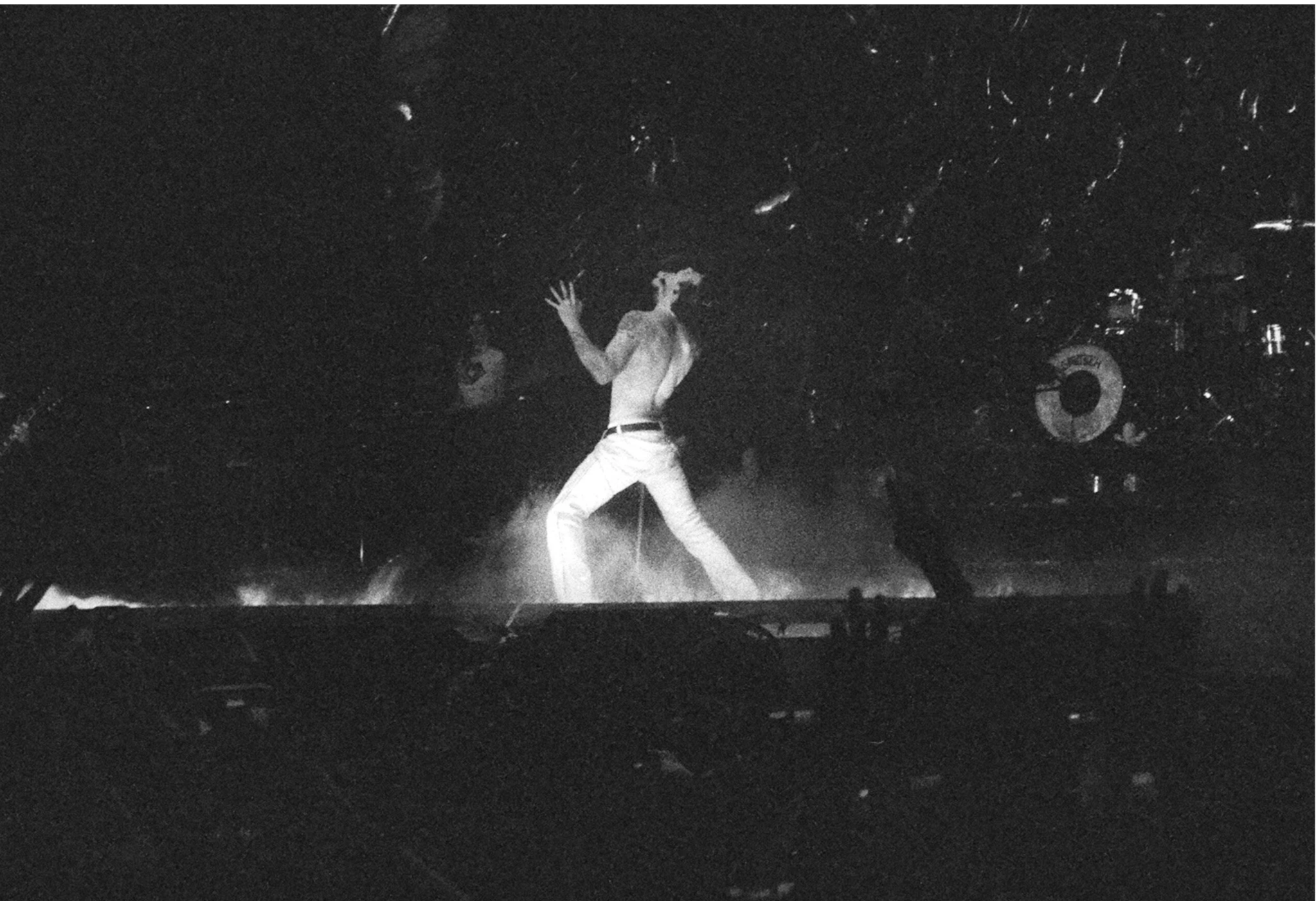 Black and white photo of a Yungblud, dressed in light-colored pants, shirtless, with one arm raised, under a dark background with some lighting effects at the O2