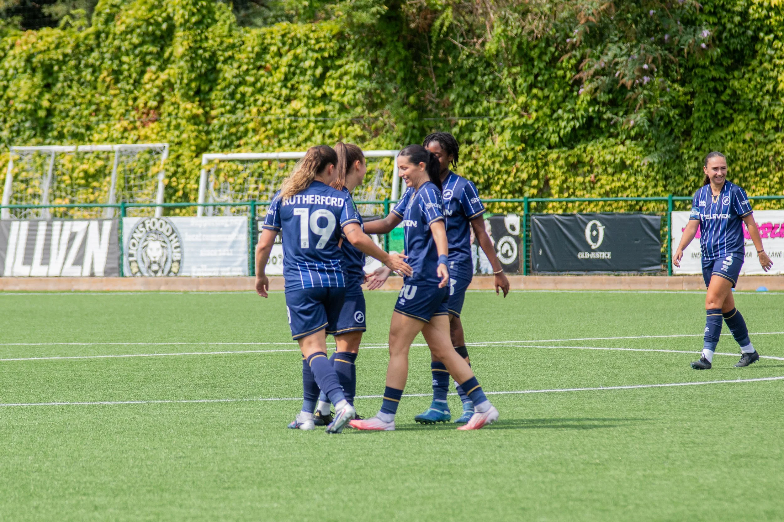 Women football players celebrating on a field, wearing blue jerseys, with greenery and sponsor banners in the background. Millwall Lionesses