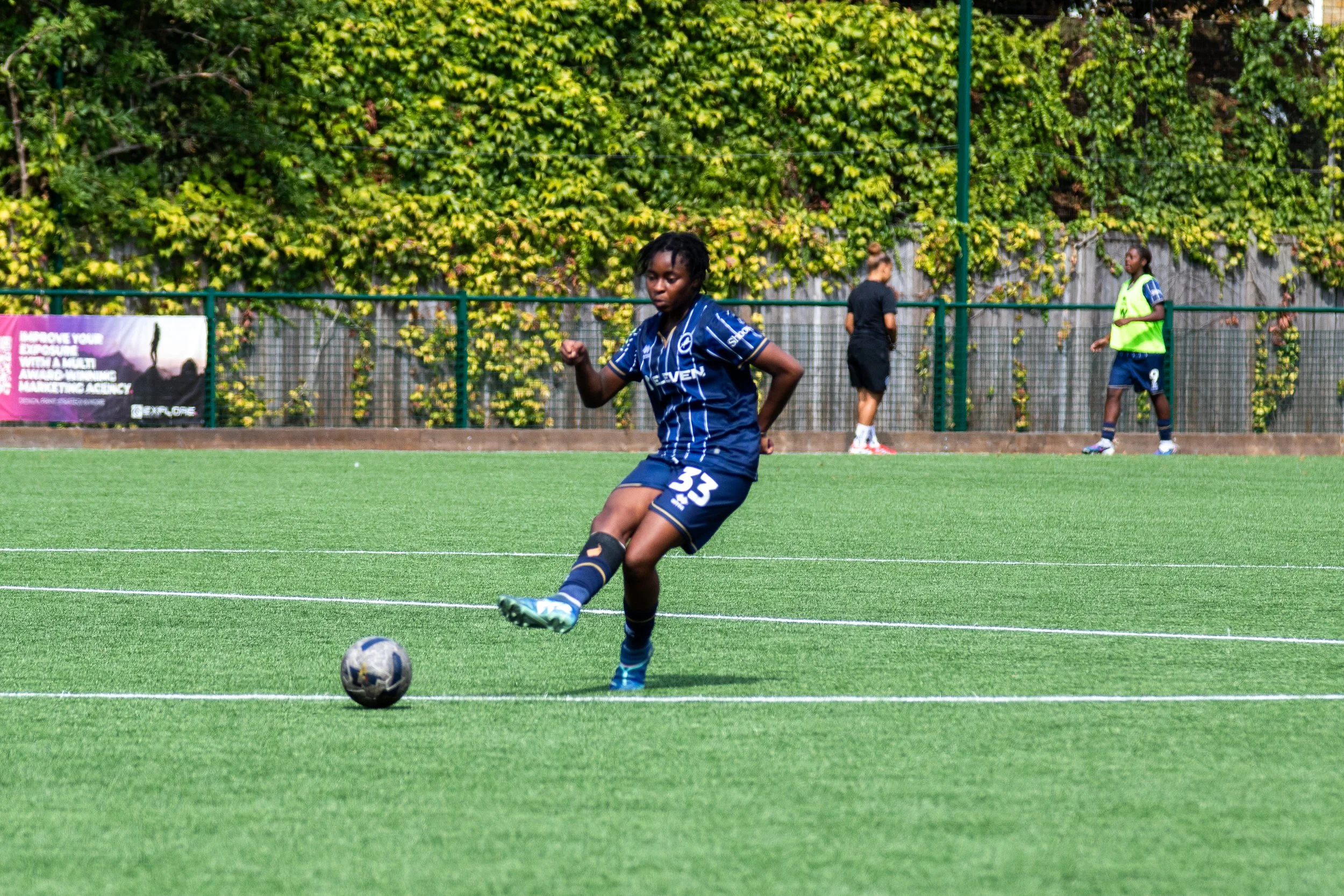 A female football player in a blue uniform with the number 33 is kicking a soccer ball on a green field. Two other players and a person in black are in the background near a fence covered with green foliage. Millwall Lionesses