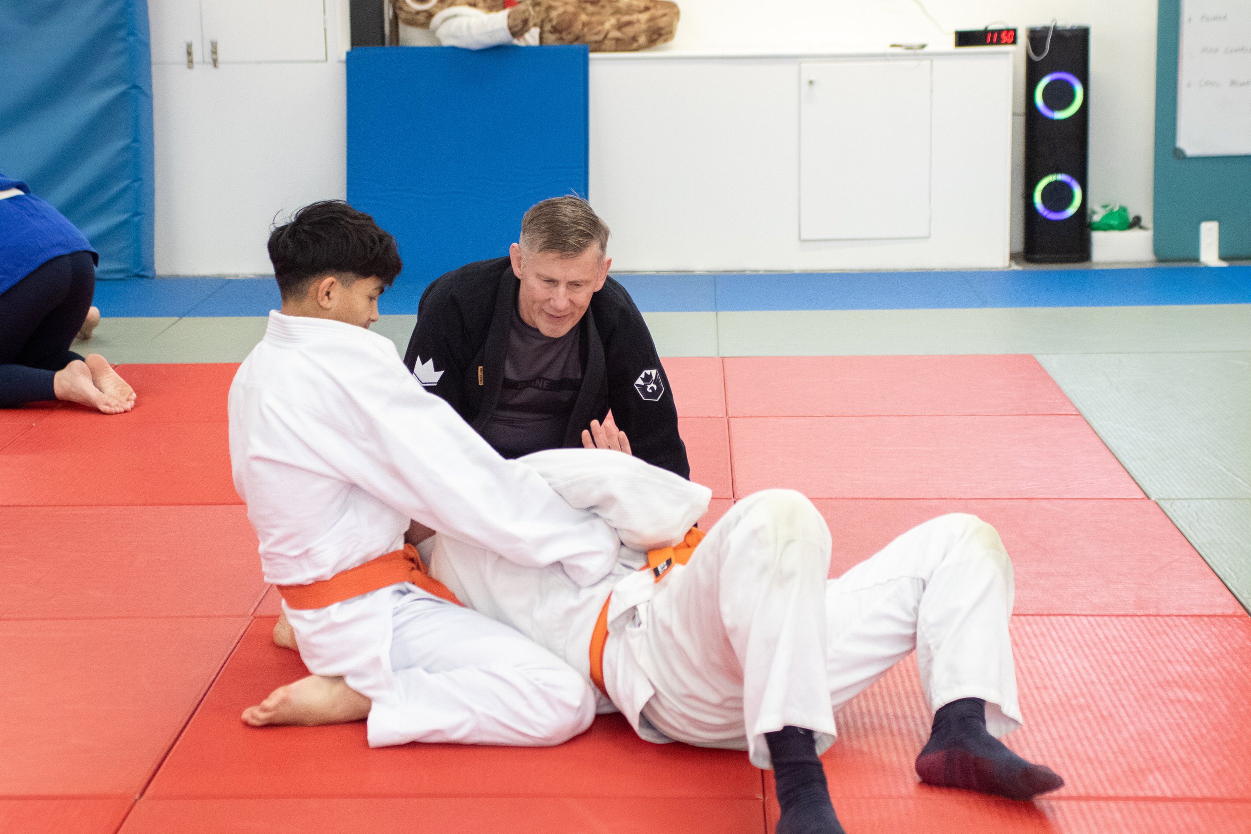 A martial arts class showing a young boy and an instructor sitting on a red mat, with others partially visible around them, in a dojo with padded blue walls and equipment, and electronic devices in the background. Relentless Judo, British Judo