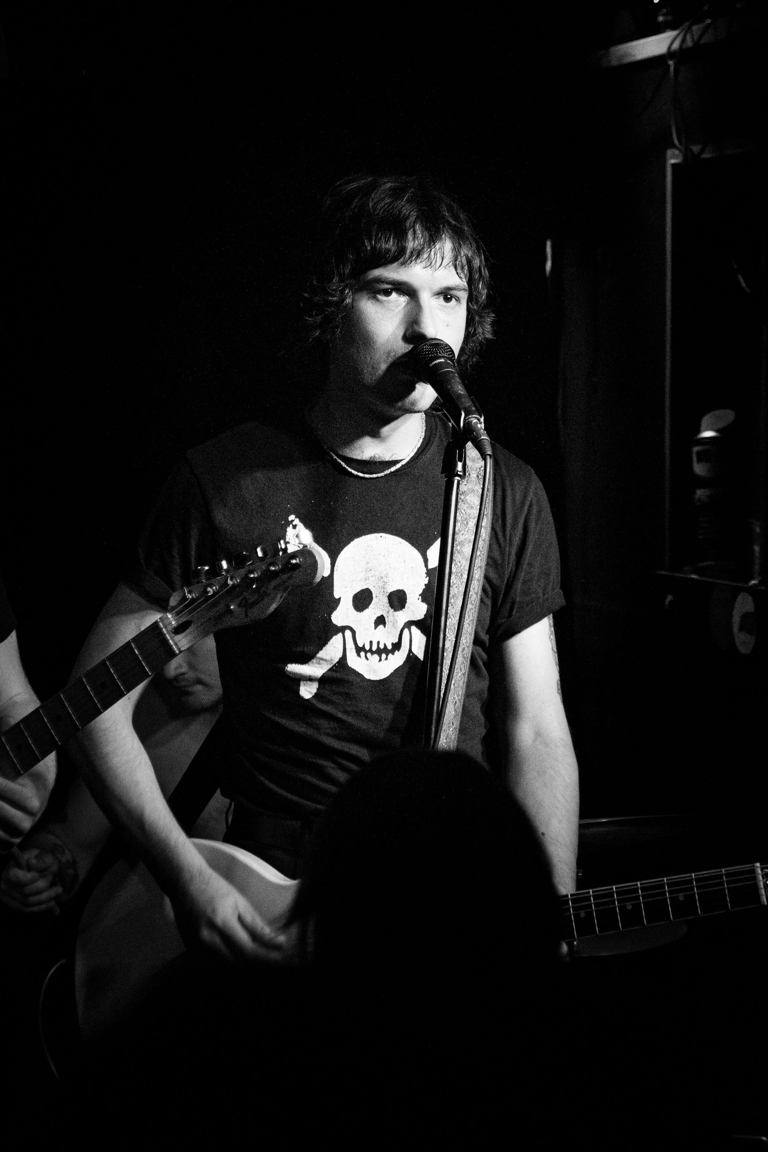 A young man with dark, curly hair sings into a microphone while playing an acoustic guitar, wearing a T-shirt with a skull and crossbones design. The scene is dimly lit, likely during a live performance in a dark music venue.