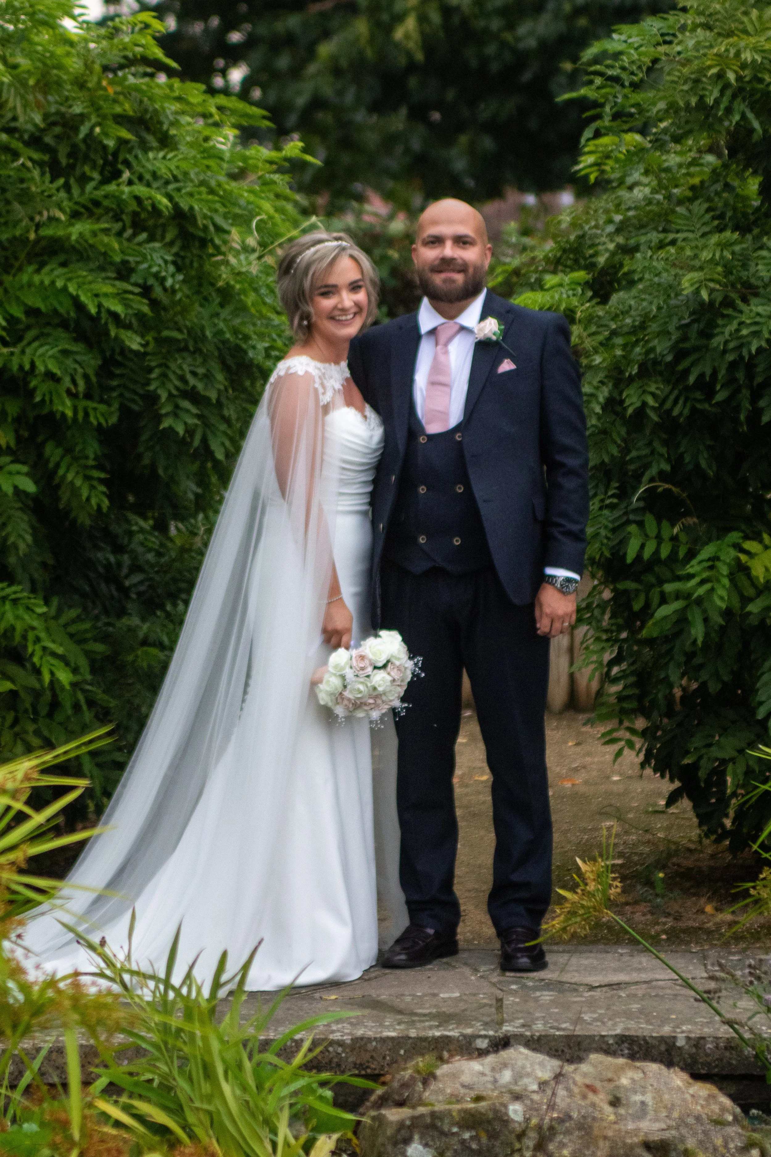 A bride and groom standing outdoors in a lush, green garden, smiling at the camera on their wedding day. The bride is wearing a white wedding dress with lace details and holding a bouquet of white and blush roses. The groom is dressed in a navy suit with a pink tie and a boutonniere, and has a beard.