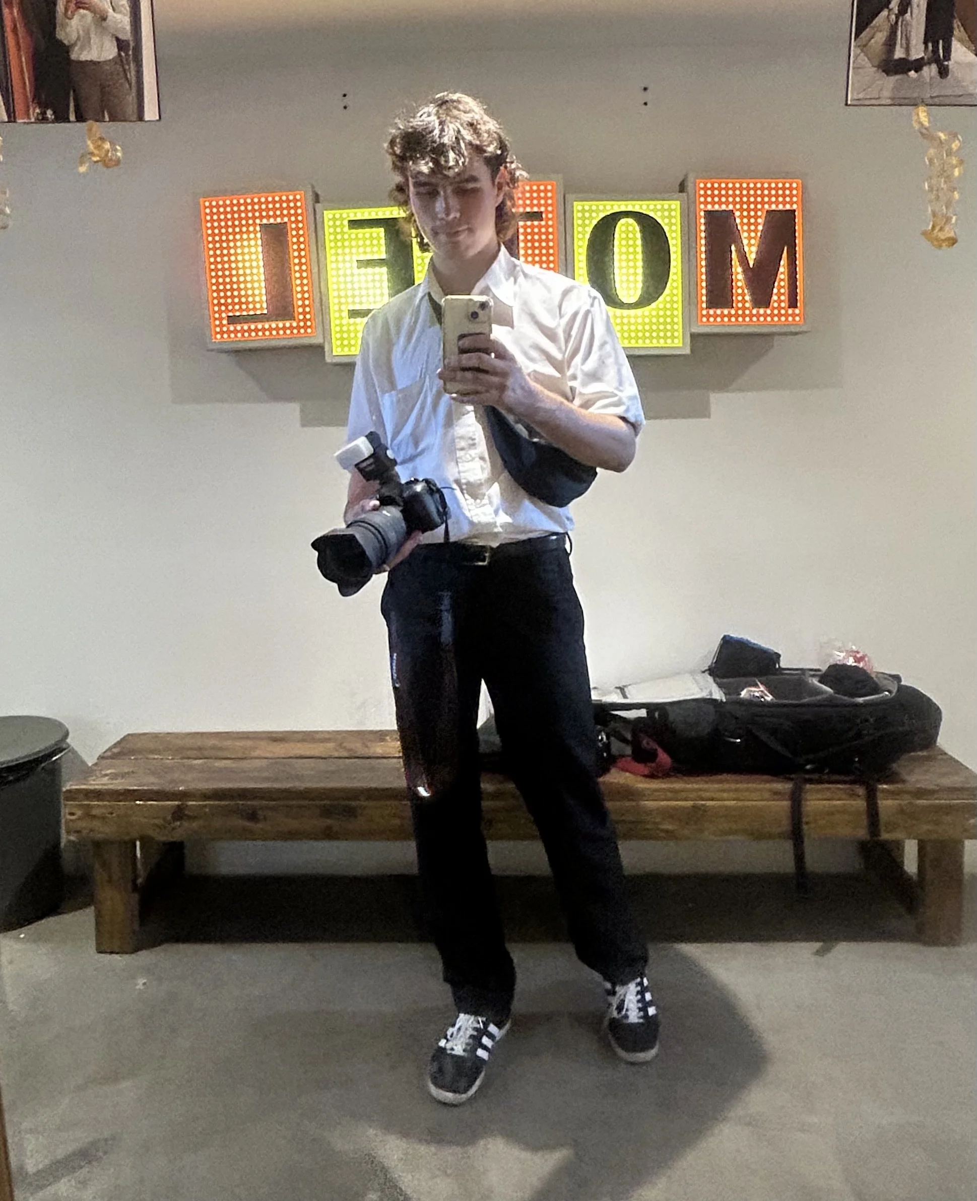 A young man with curly hair taking a selfie in front of a colorful illuminated sign that spells 'WELCOME'. He is wearing a white shirt, black pants, and black sneakers with white laces, and is holding a camera in his left hand. There is a camera bag and other items on a wooden table behind him, with a gray floor and a gray wall in the background.