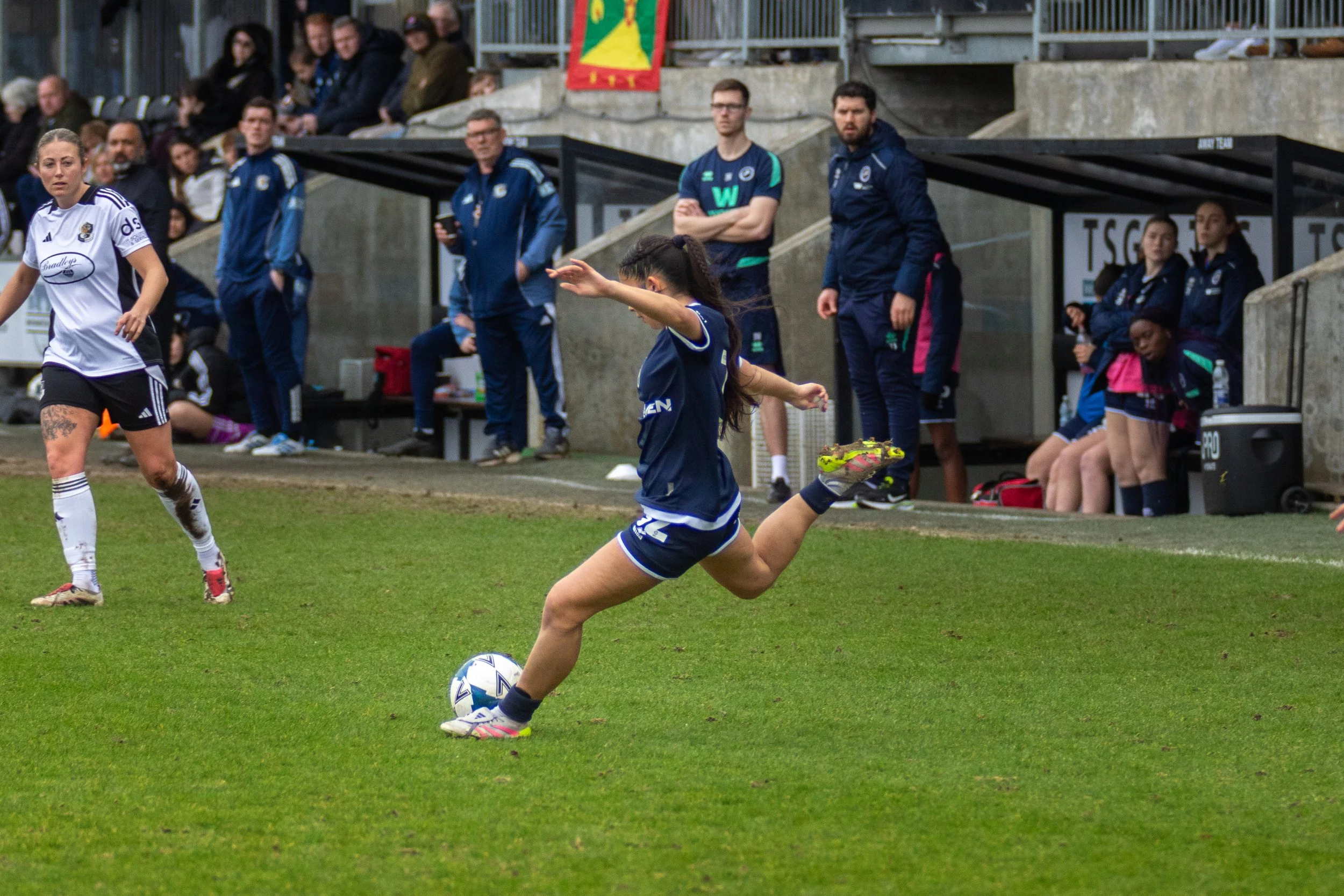 A woman in a blue football uniform kicking a ball on a football pitch during a match, with spectators and team staff observing from the bench area. Millwall Lionesses