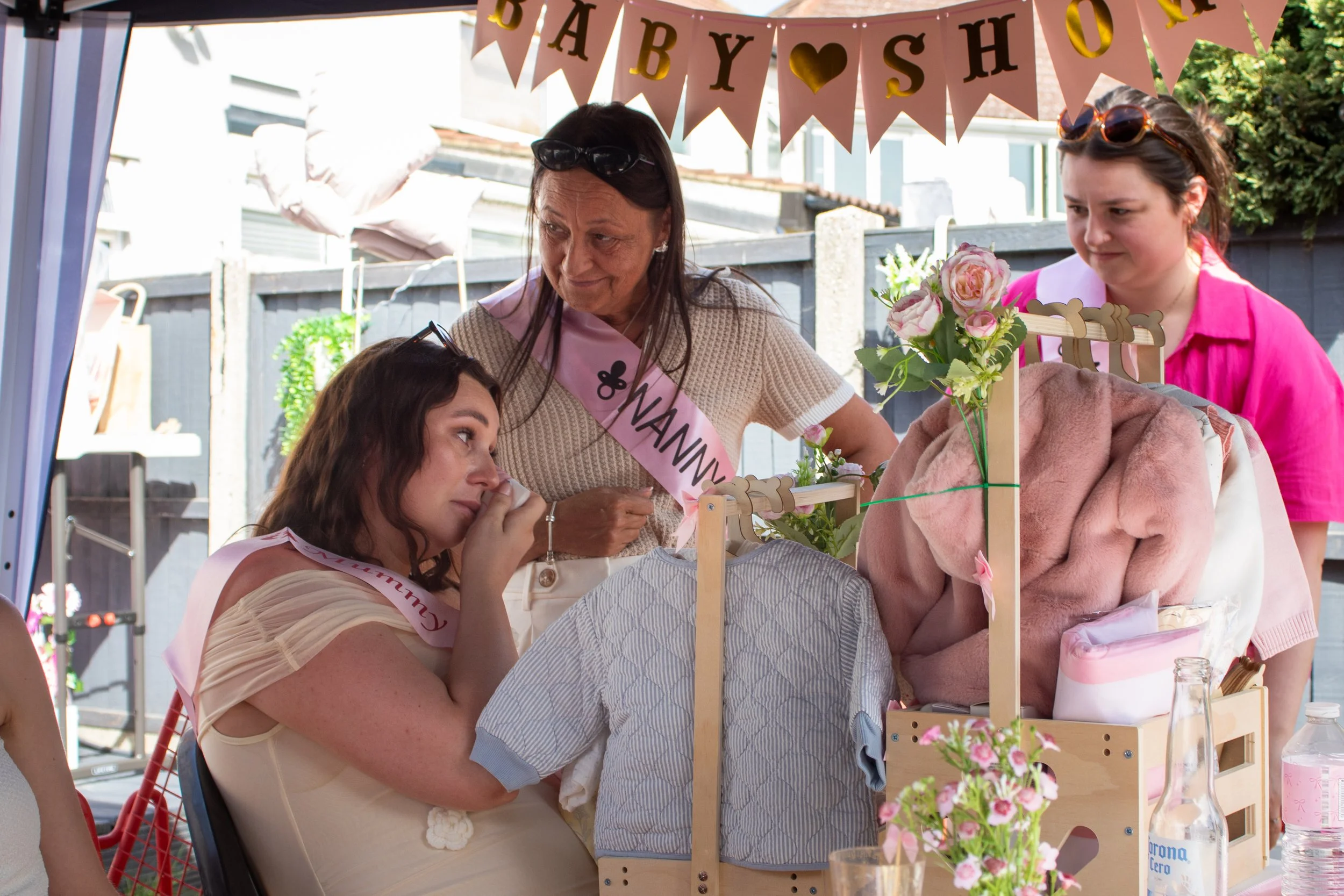 A baby shower celebration with women gathered around a table outdoors. Decorations include pink banners spelling 'BABY SHOWER' with gold hearts. A woman with a pink sash and sunglasses on her head is looking at a baby in a basket, surrounded by flowe