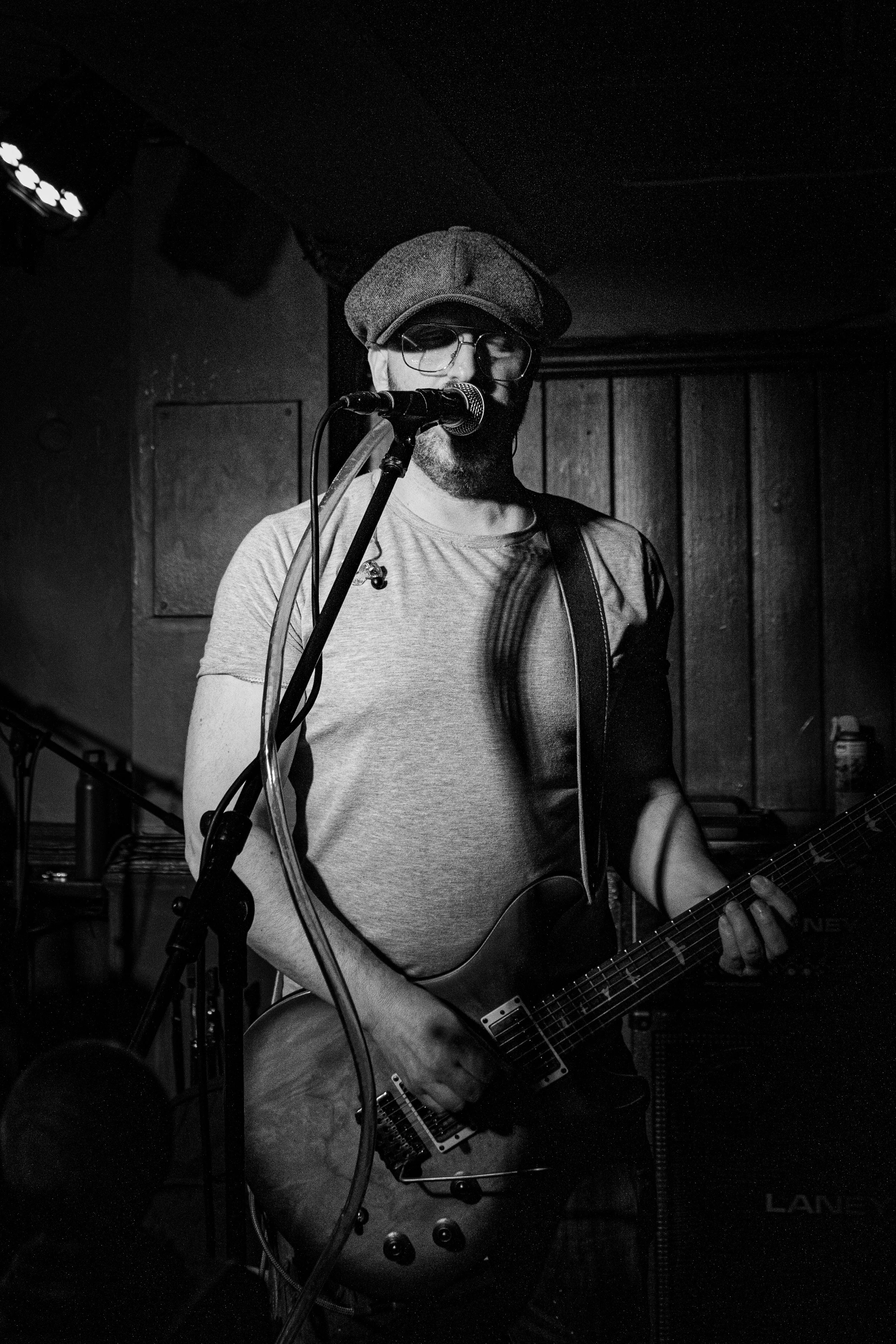A man wearing glasses and a flat cap playing an acoustic guitar and singing into a microphone at a dimly lit venue.