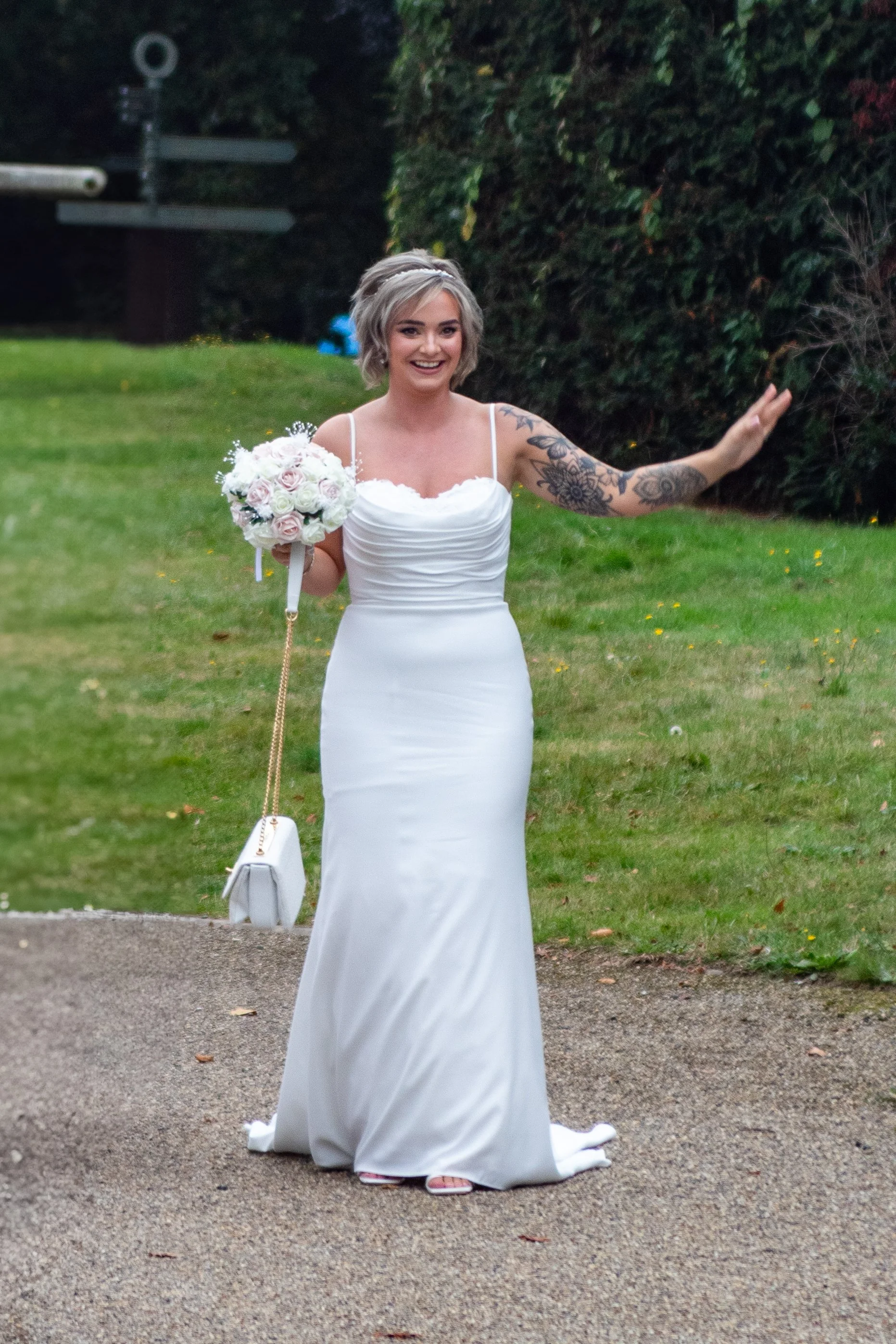 A woman is standing outdoors on a gravel path surrounded by grass and trees, wearing a white wedding dress and holding a bouquet of flowers.