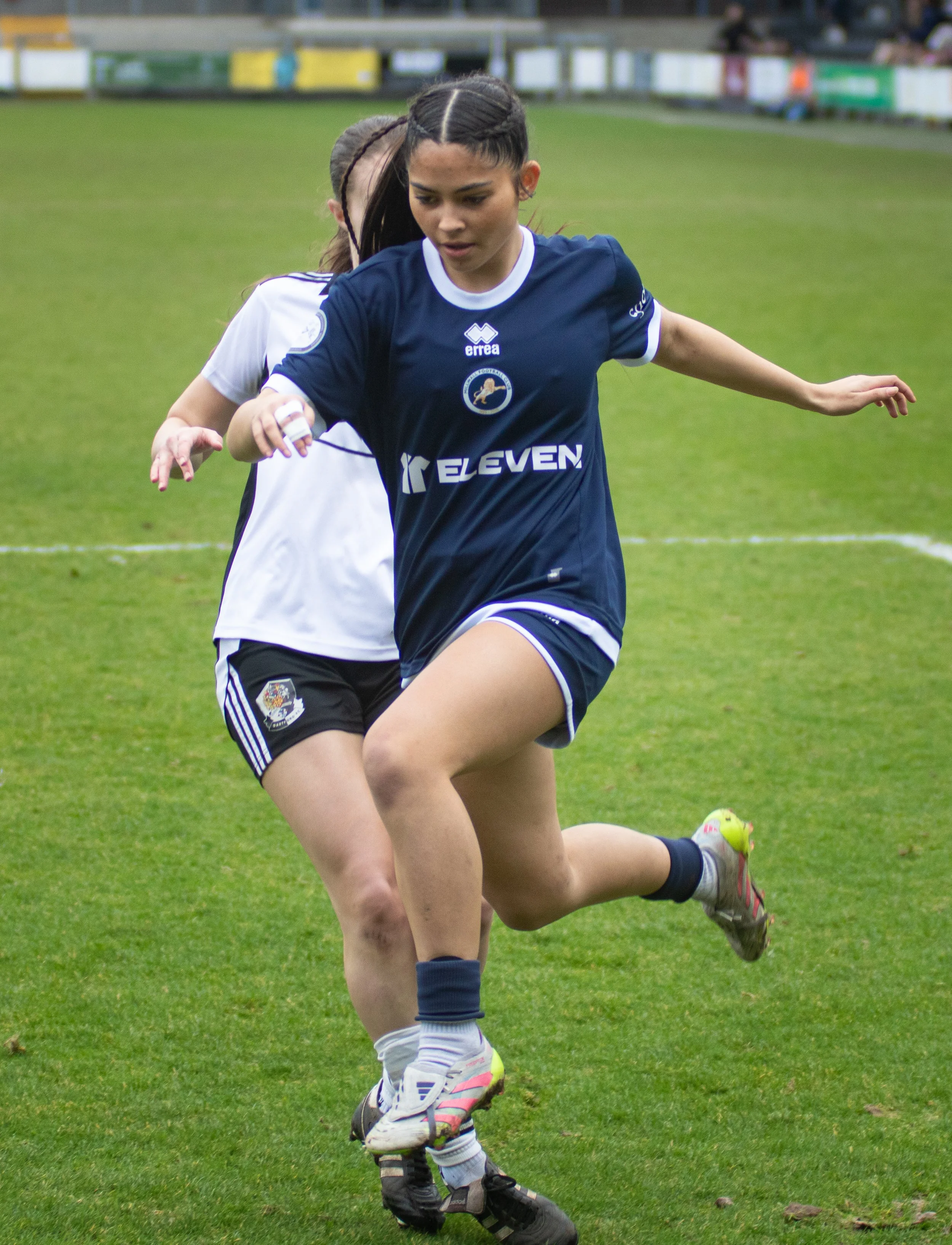 Two female football players competing for the ball on a grassy field, with one wearing a dark blue jersey and the other in a white jersey, in a stadium setting. Millwall Lionesses