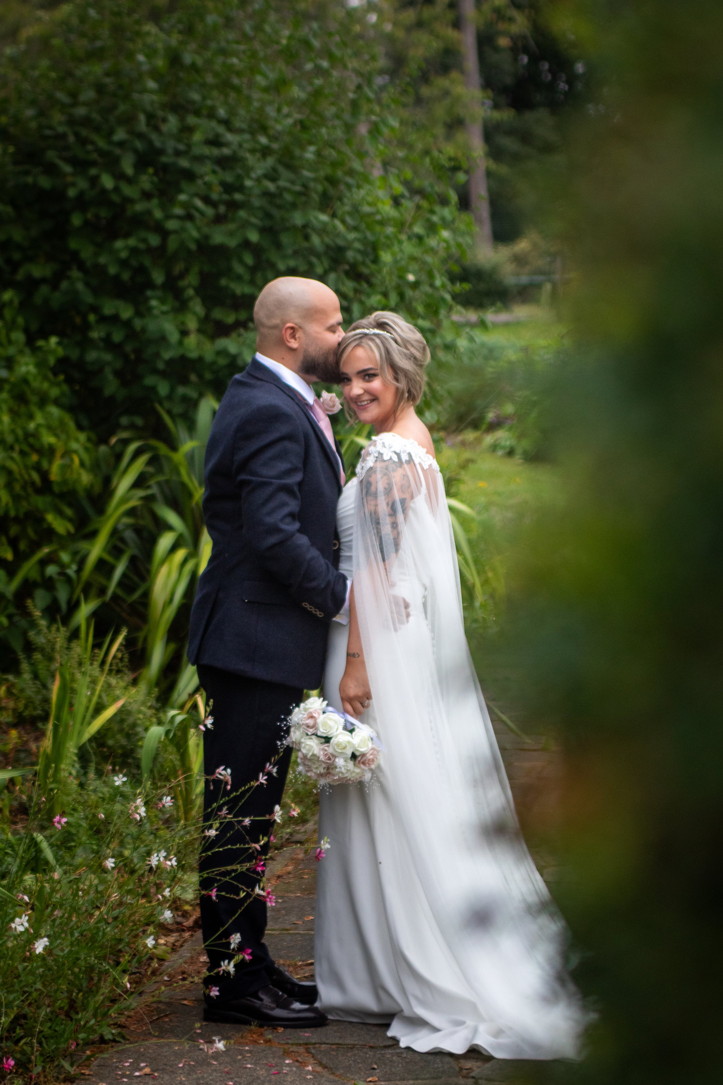A newlywed couple standing on a garden pathway surrounded by green foliage. The bride is smiling and holding a bouquet of white roses, wearing a white wedding gown with lace sleeves and a veil. The groom, bald with a beard, is kissing her on the head