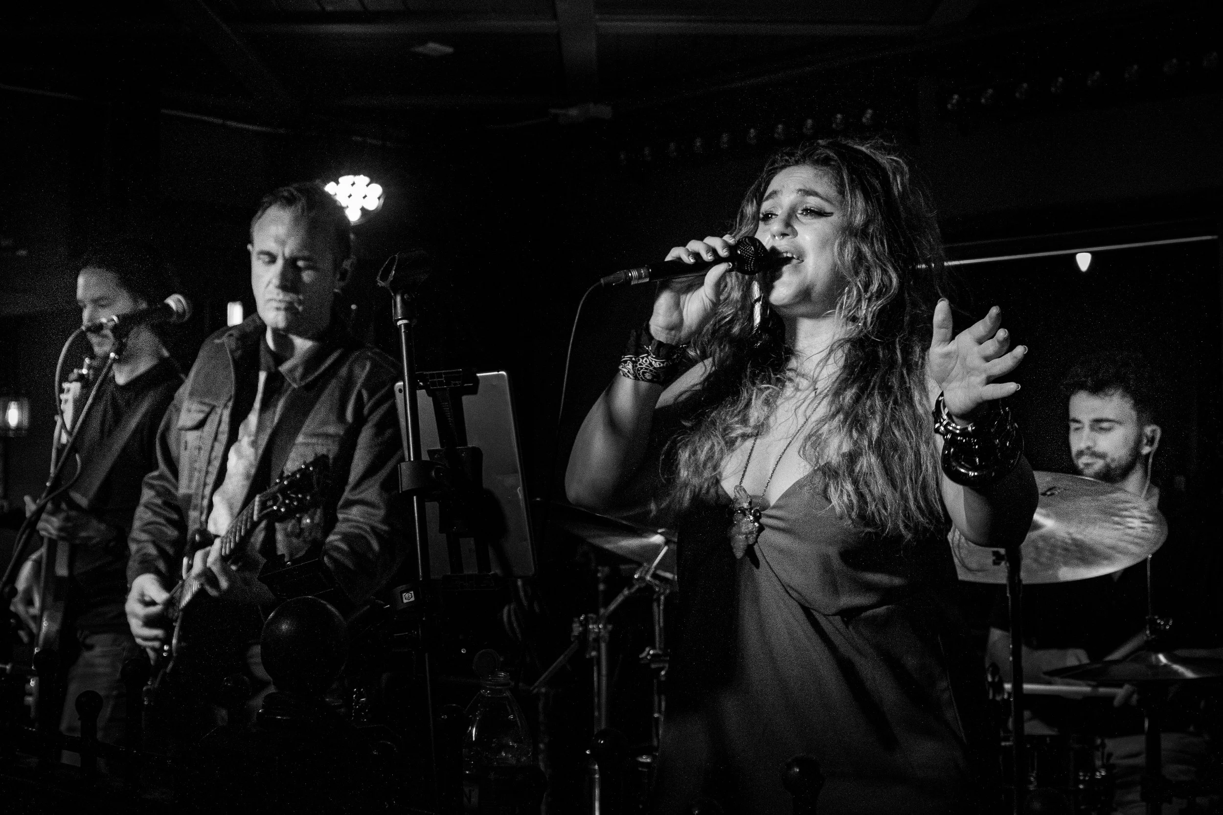 A black and white photograph of a female singer with long, wavy hair singing into a microphone on stage. She is wearing a strapless dress and accessories, with her hand raised. Behind her are band members playing instruments, including a guitarist an
