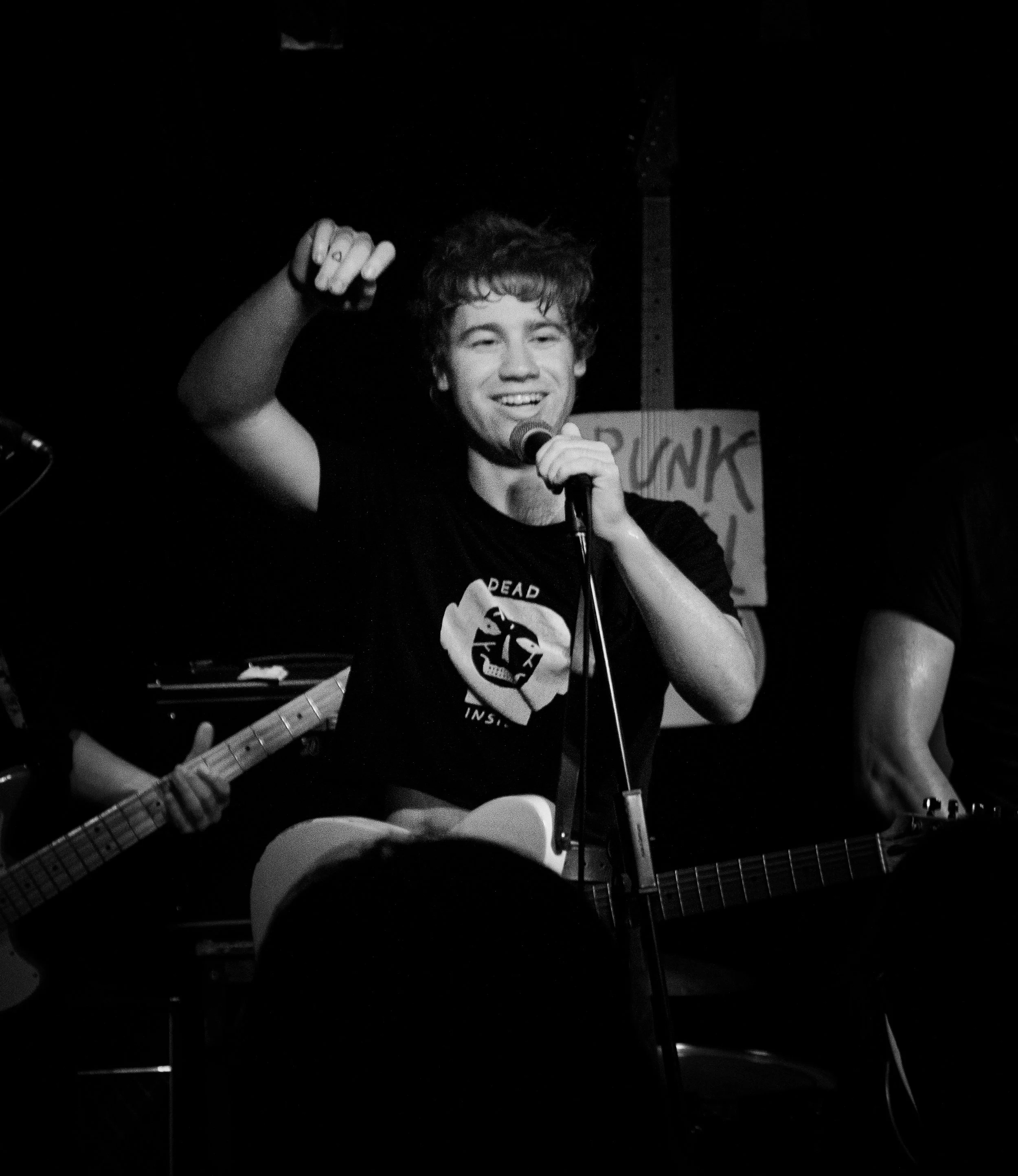 Young man with curly hair and a big smile singing into a microphone while playing an acoustic guitar at a live music event. A hand with a guitar neck is visible in the foreground. The background is dark with some signs and equipment.