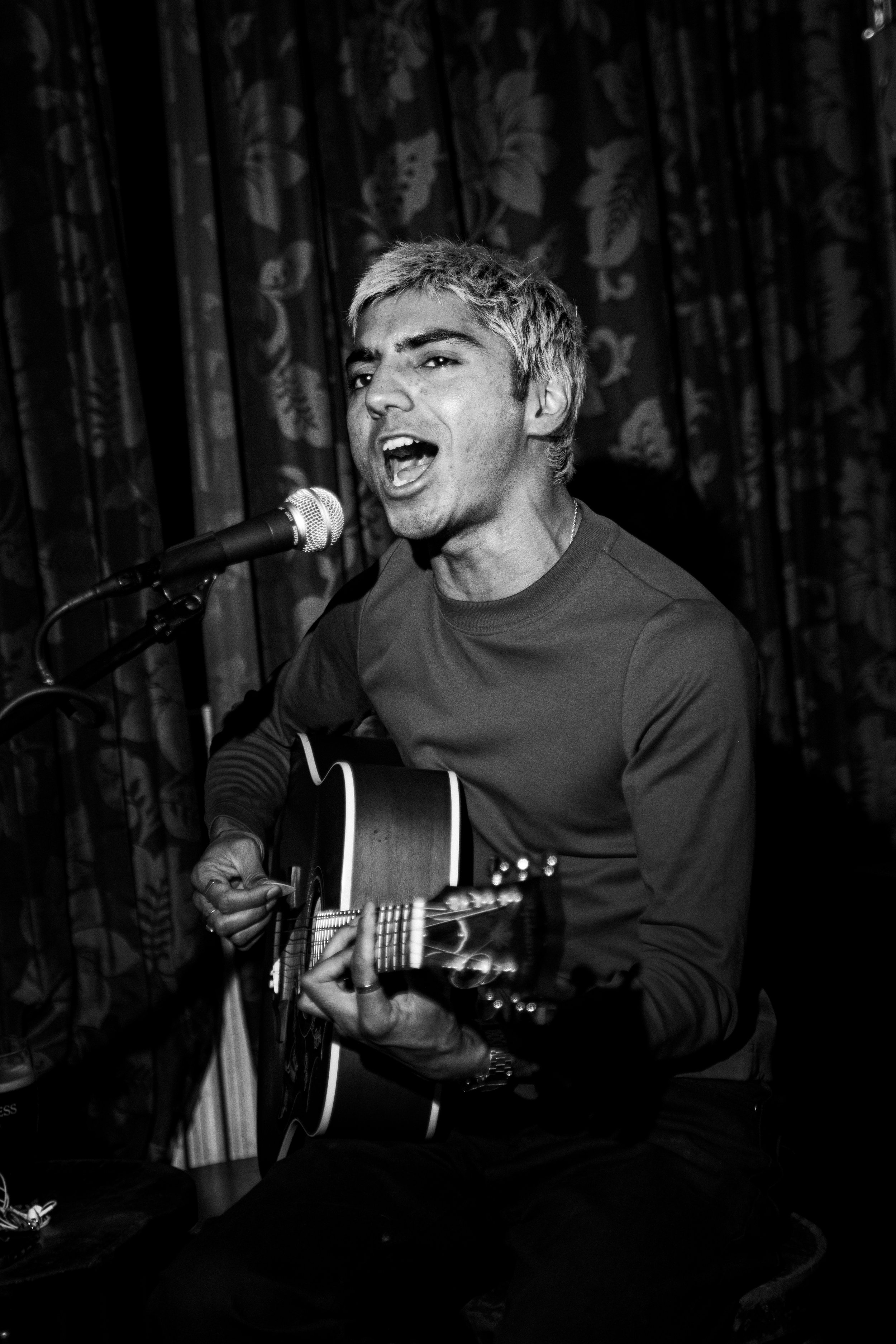 A black and white photo of a man singing into a microphone while playing an acoustic guitar, with a decorative curtain in the background. The Hawley Arms, Camden