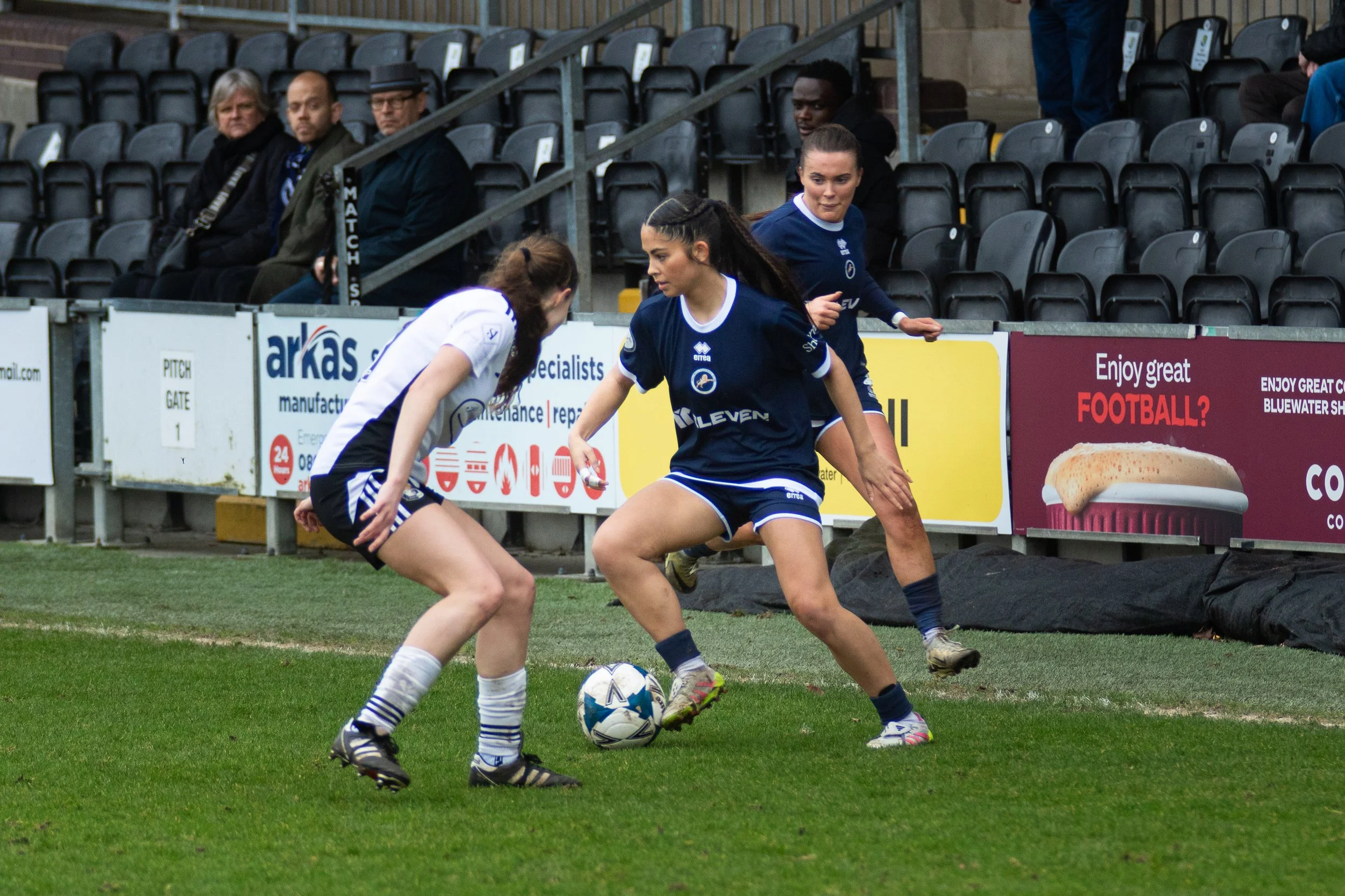 Women playing football on the field, with some spectators sitting in the stands in the background. Millwall Lionesses