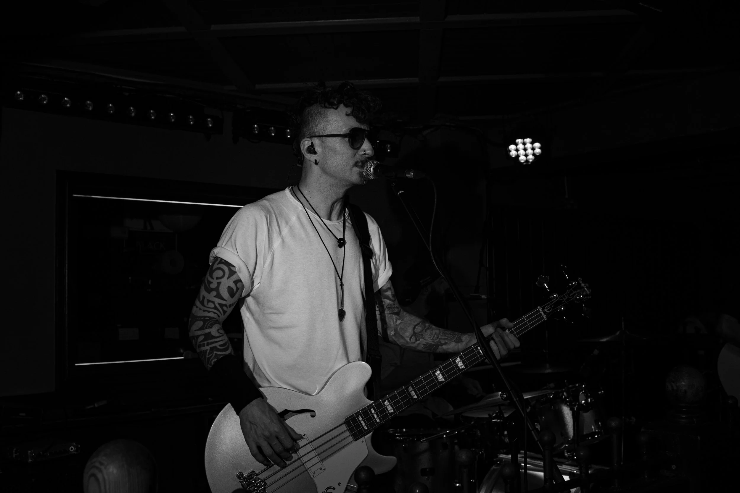 A musician with tattoos on his arm, wearing glasses and a white shirt, playing an electric bass guitar and singing into a microphone on a dark stage with stage lights.