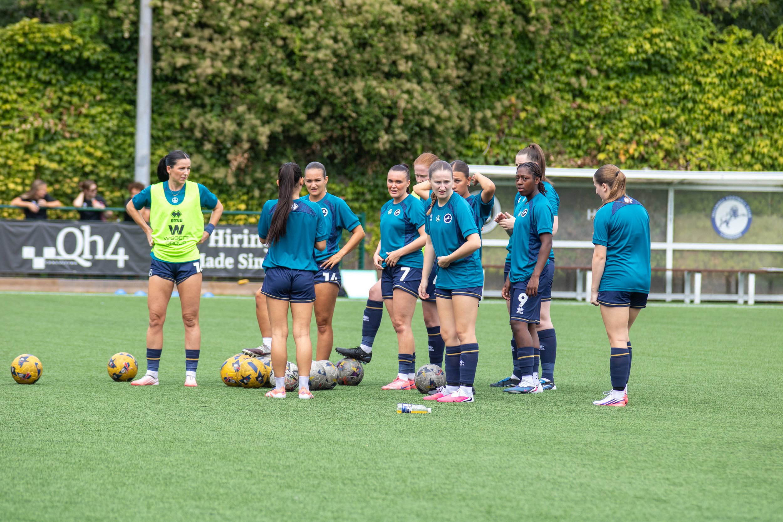 Group of female football players standing on the field during a practice or team meeting, with footballs balls lined up in front of them. Millwall Lionesses