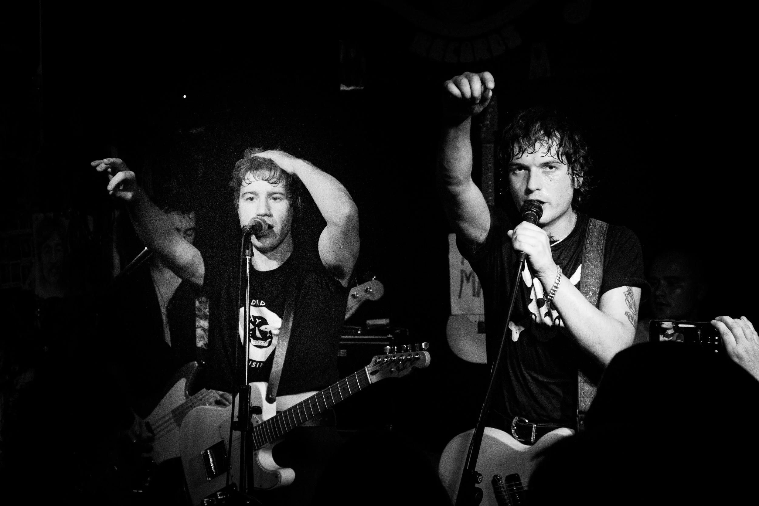 Two men with guitars singing into microphones on a dark stage, raising fists, with a background banner and musical equipment.