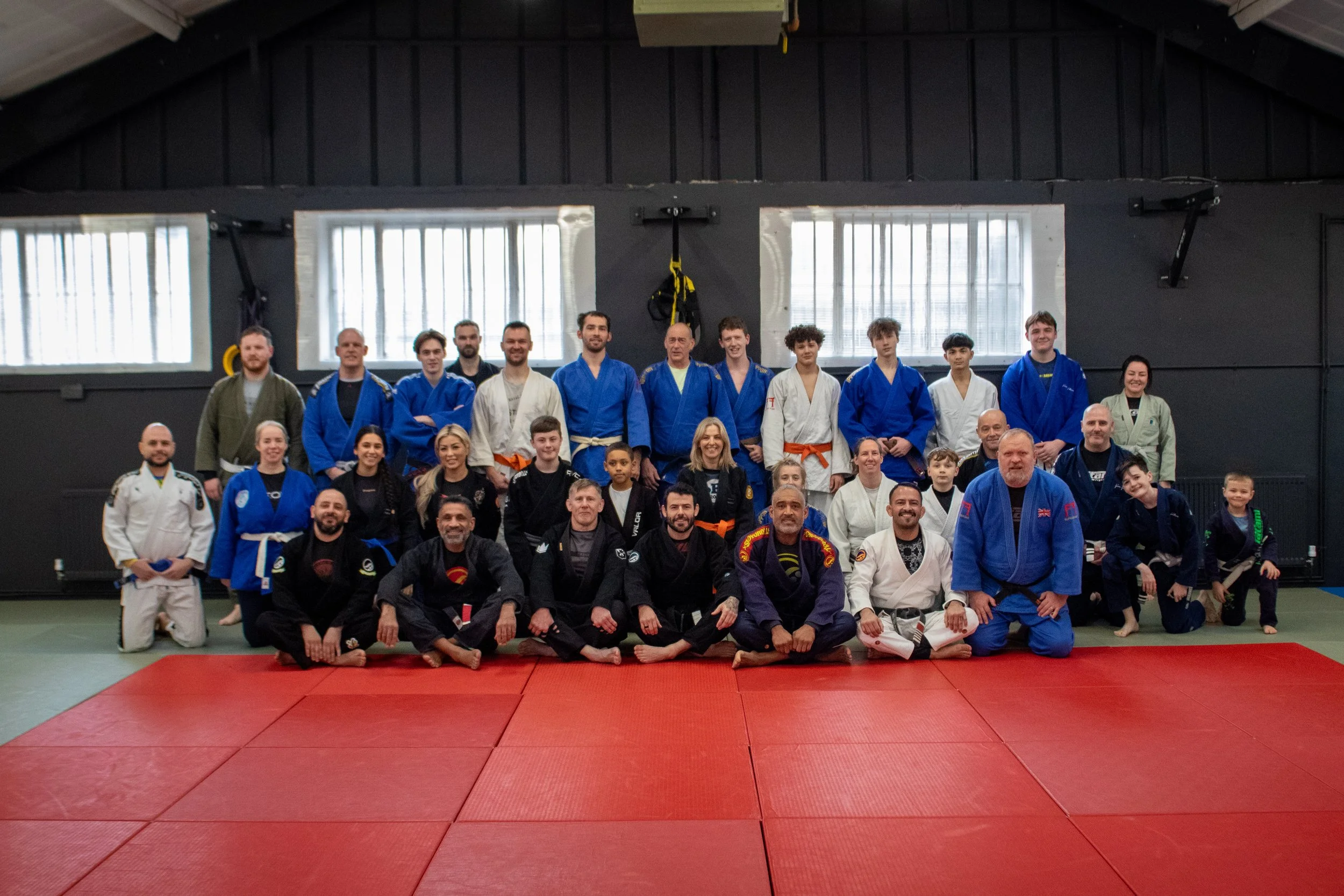 Group of adults and children in martial arts uniforms posing on mats in a dojo or training gym. Relentless Judo, British Judo