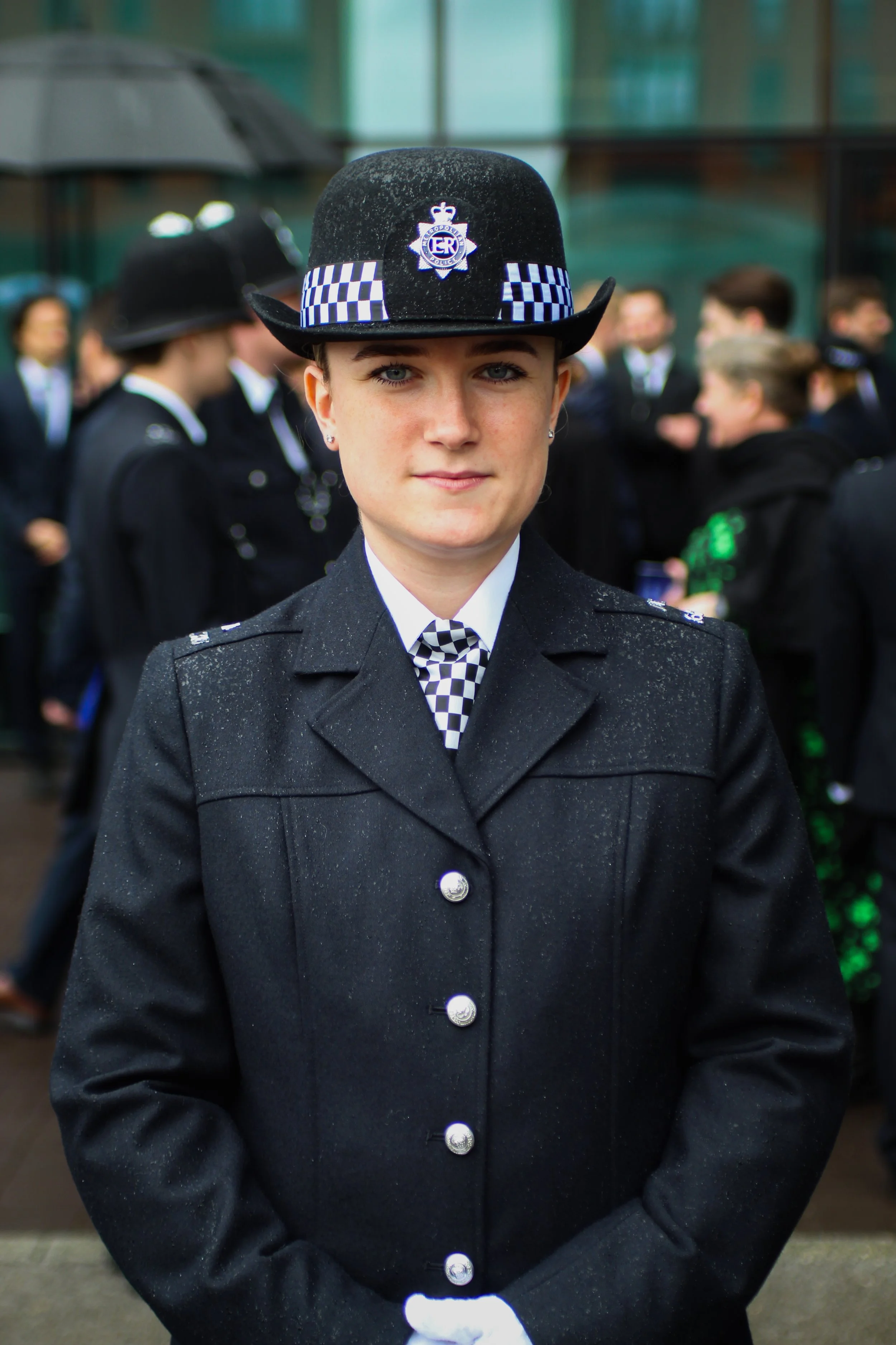 A female police officer wearing a formal uniform and a traditional custodian helmet with a badge, standing outdoors in a group of people.